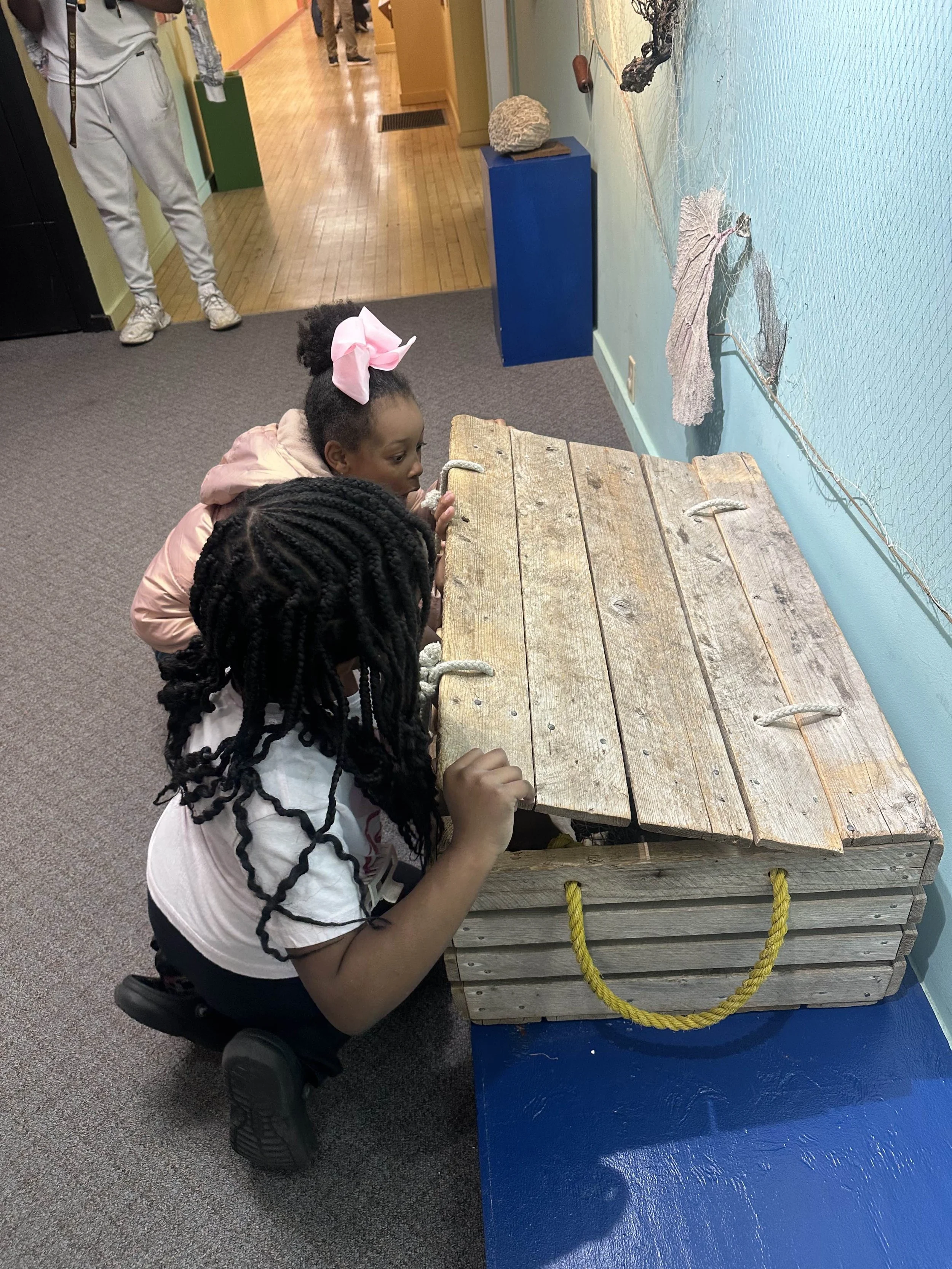 Two young girls are kneeling and leaning over a wooden cargo net or trap at a museum or educational exhibit, observing something inside. One girl has black braided hair and is wearing a white t-shirt with a design, while the other girl has curly hair
