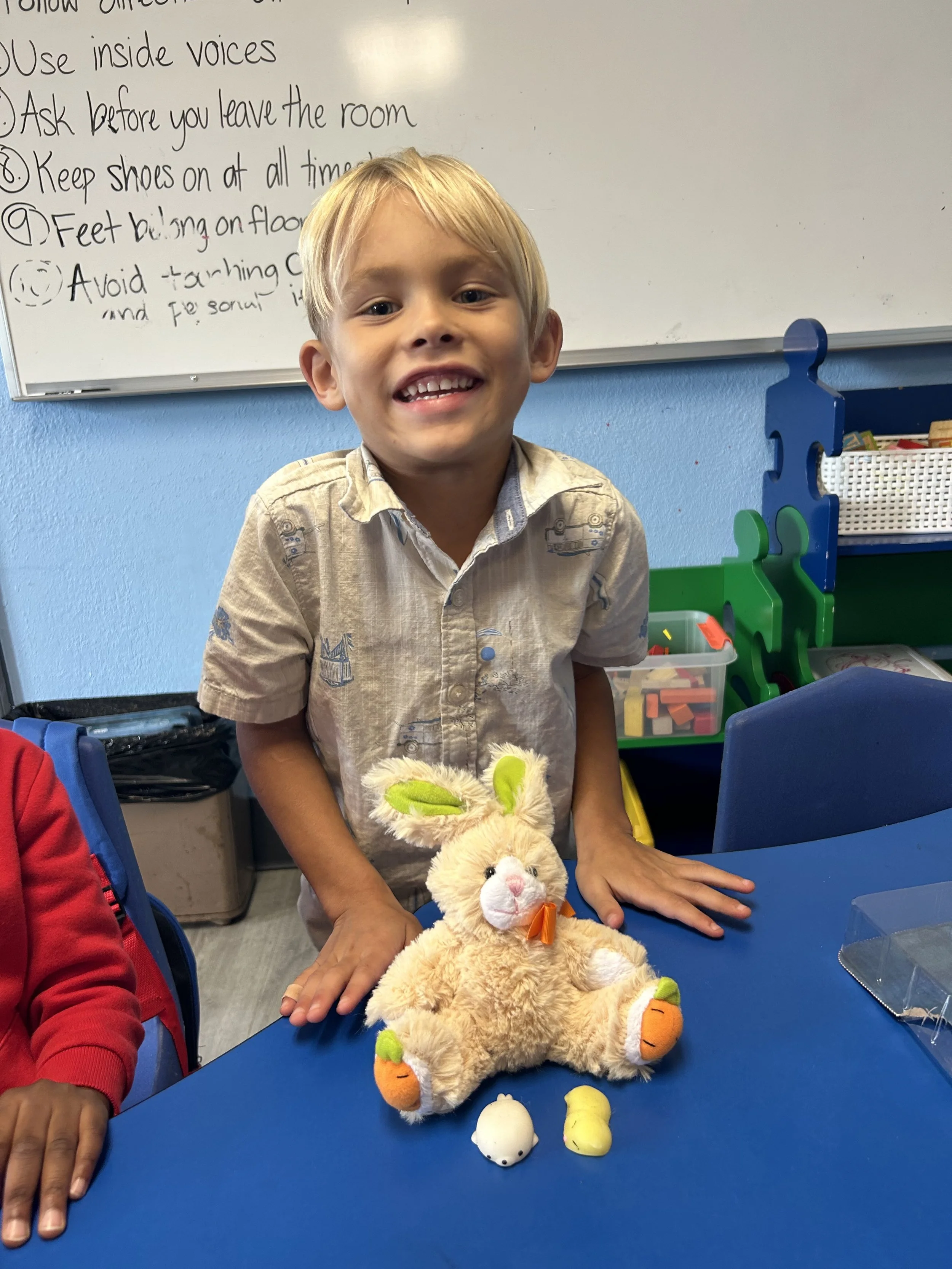 A young boy smiling and showing his teeth, standing at a blue table in a classroom. In front of him on the table is a plush bunny toy with green ear accents, orange feet, and a white face, along with two small white and yellow objects.