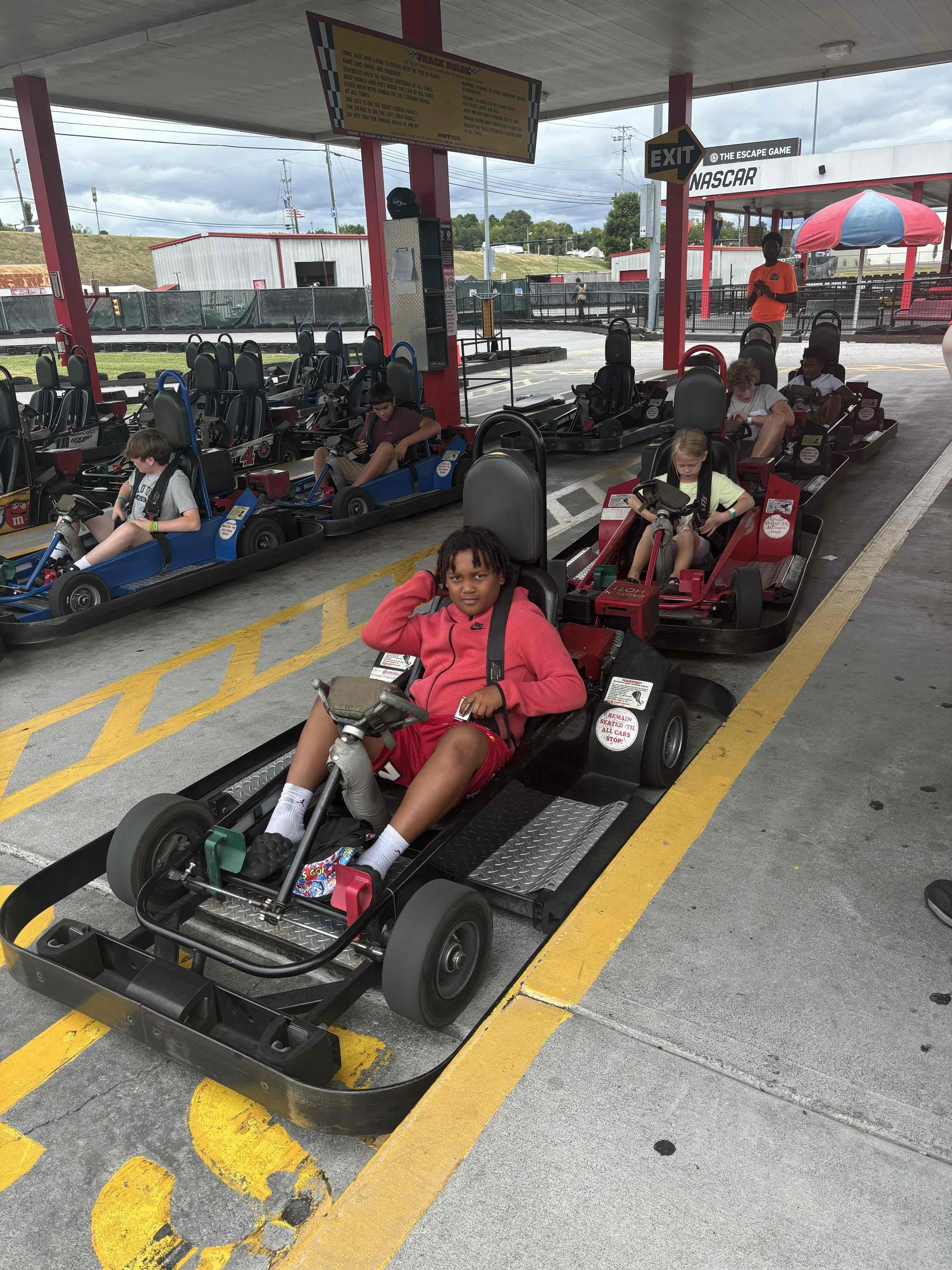 Children sitting in go-karts at an outdoor go-kart race track at NASCAR amusement park, under a shelter with a sign and exit arrow, with cloudy sky in the background.