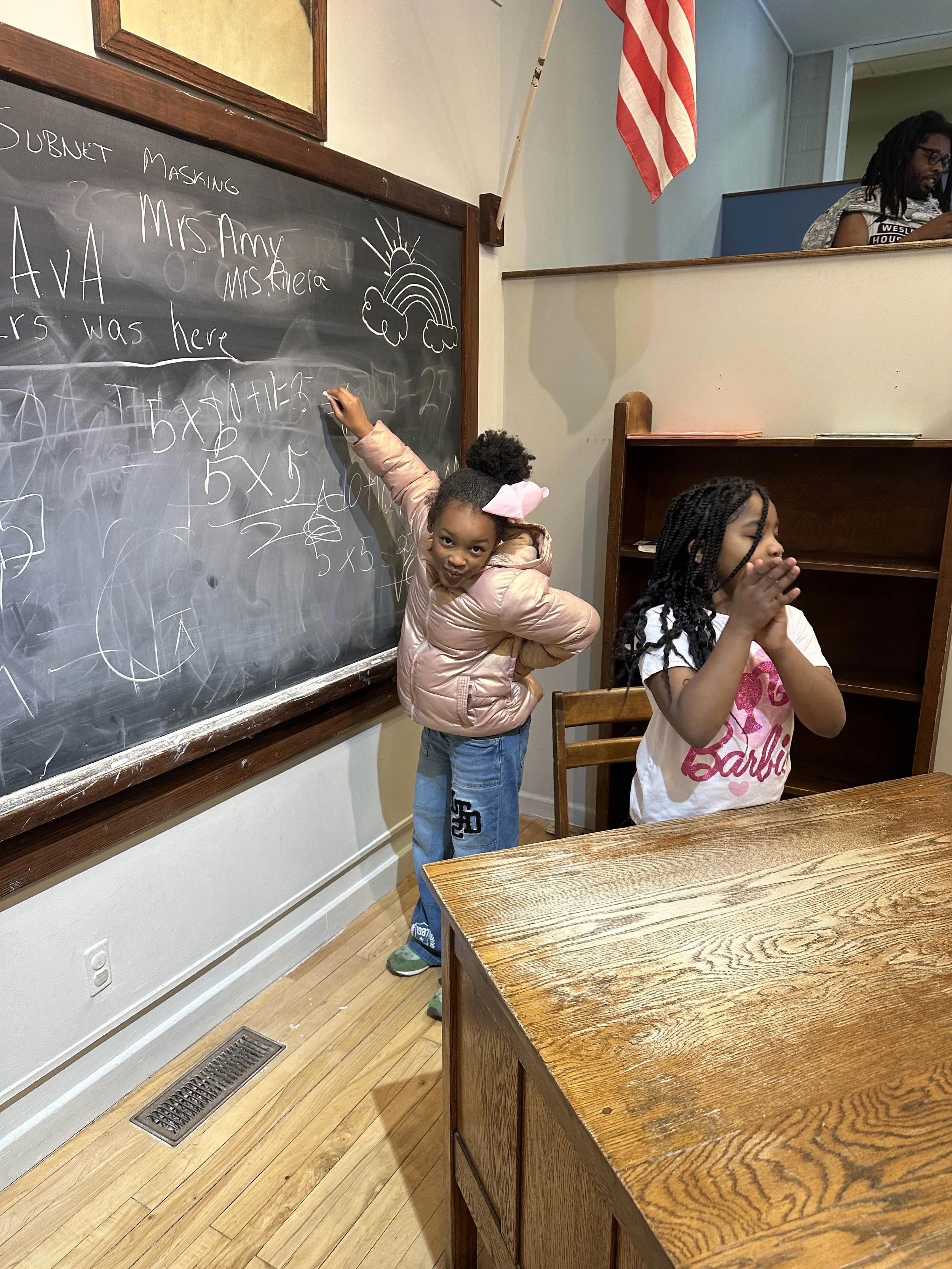 Two young girls are in a classroom near a blackboard. One girl is standing, writing on the board, wearing a pink jacket and blue jeans. The other girl is sitting at a wooden table, praying with her hands clasped, wearing a white t-shirt with a pink d