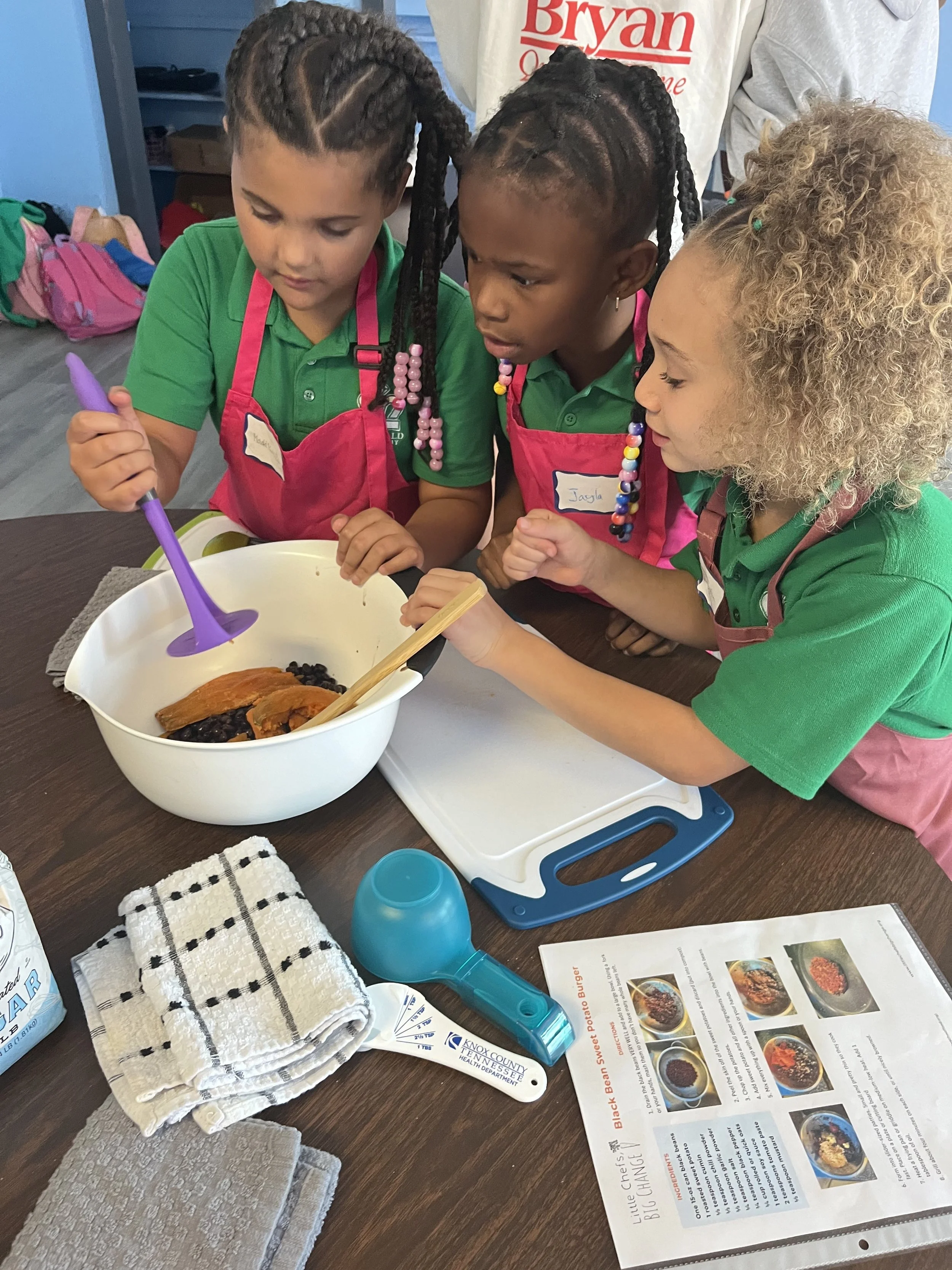 Three young girls wearing aprons and green shirts gathered around a mixing bowl with cooked black beans and sweet potatoes, participating in a cooking activity in a classroom or kitchen setting.