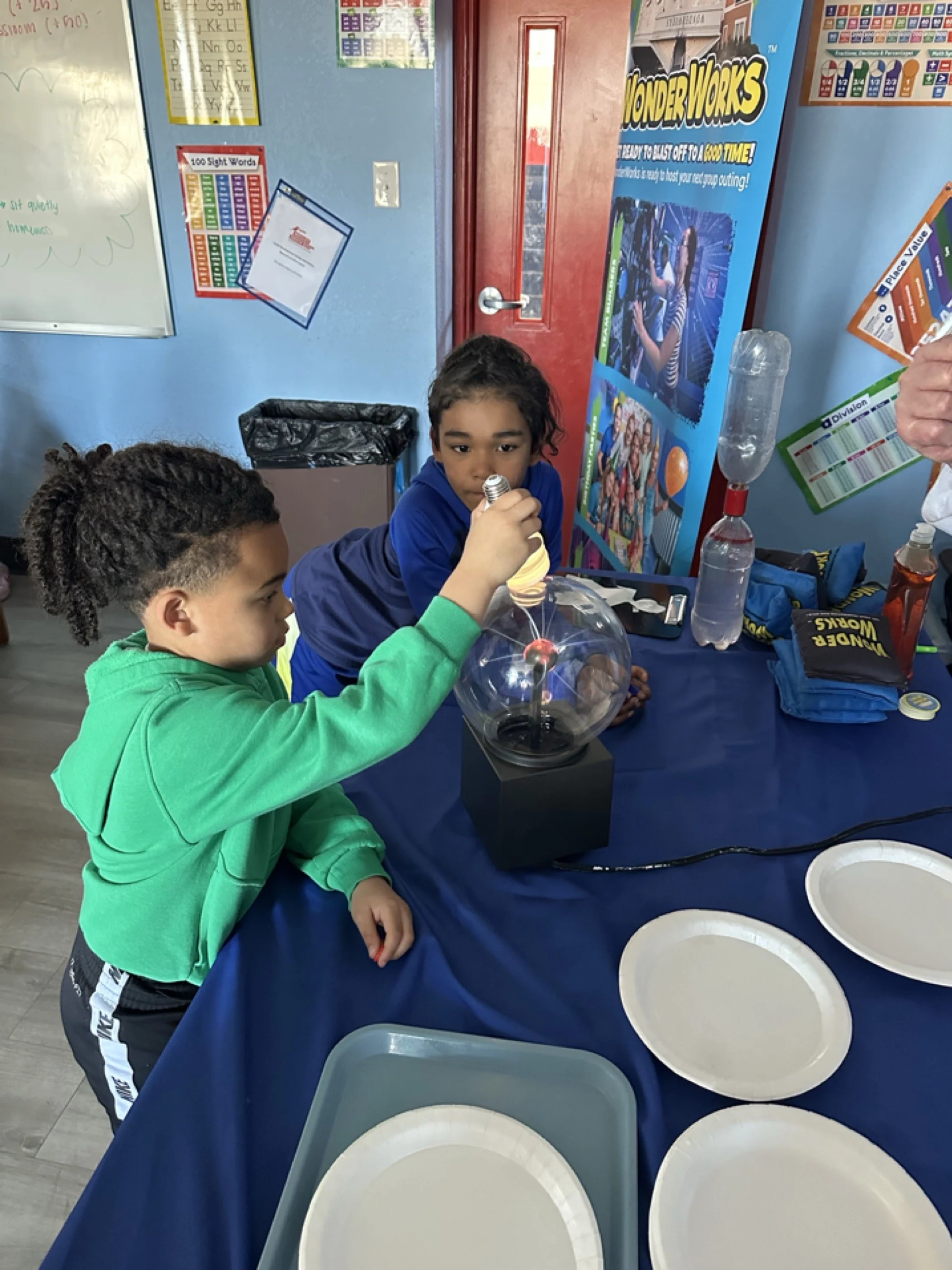 Two young girls conducting a science experiment involving a balloon and a light bulb in a classroom setting, with science posters and plates on the table.