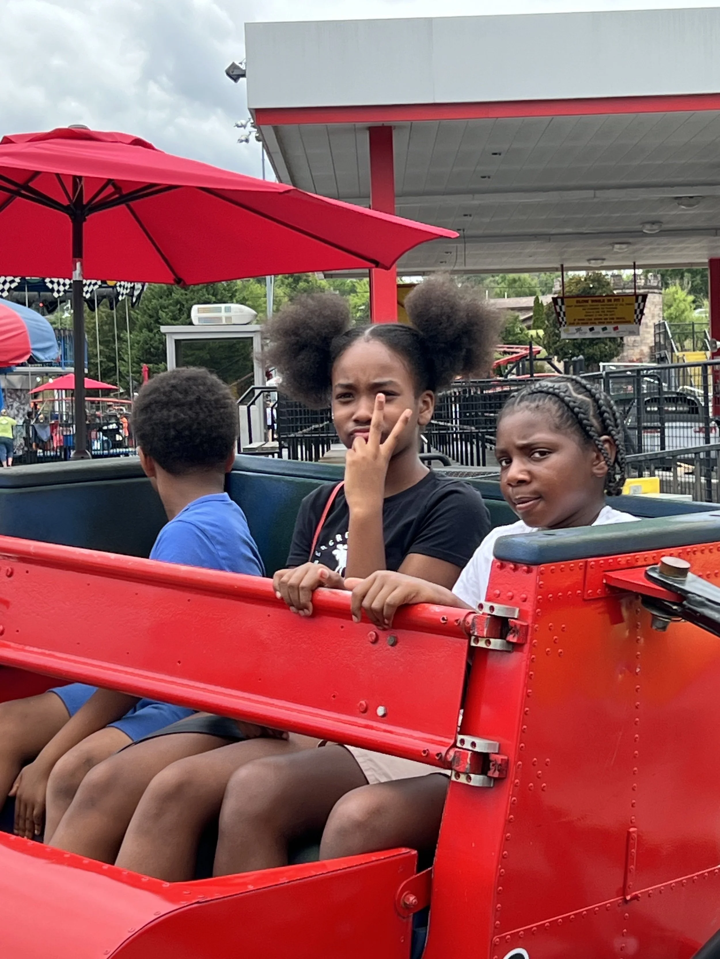Three children sitting in a red amusement park ride with a canopy, with a young girl making a peace sign and another girl sticking out her tongue.