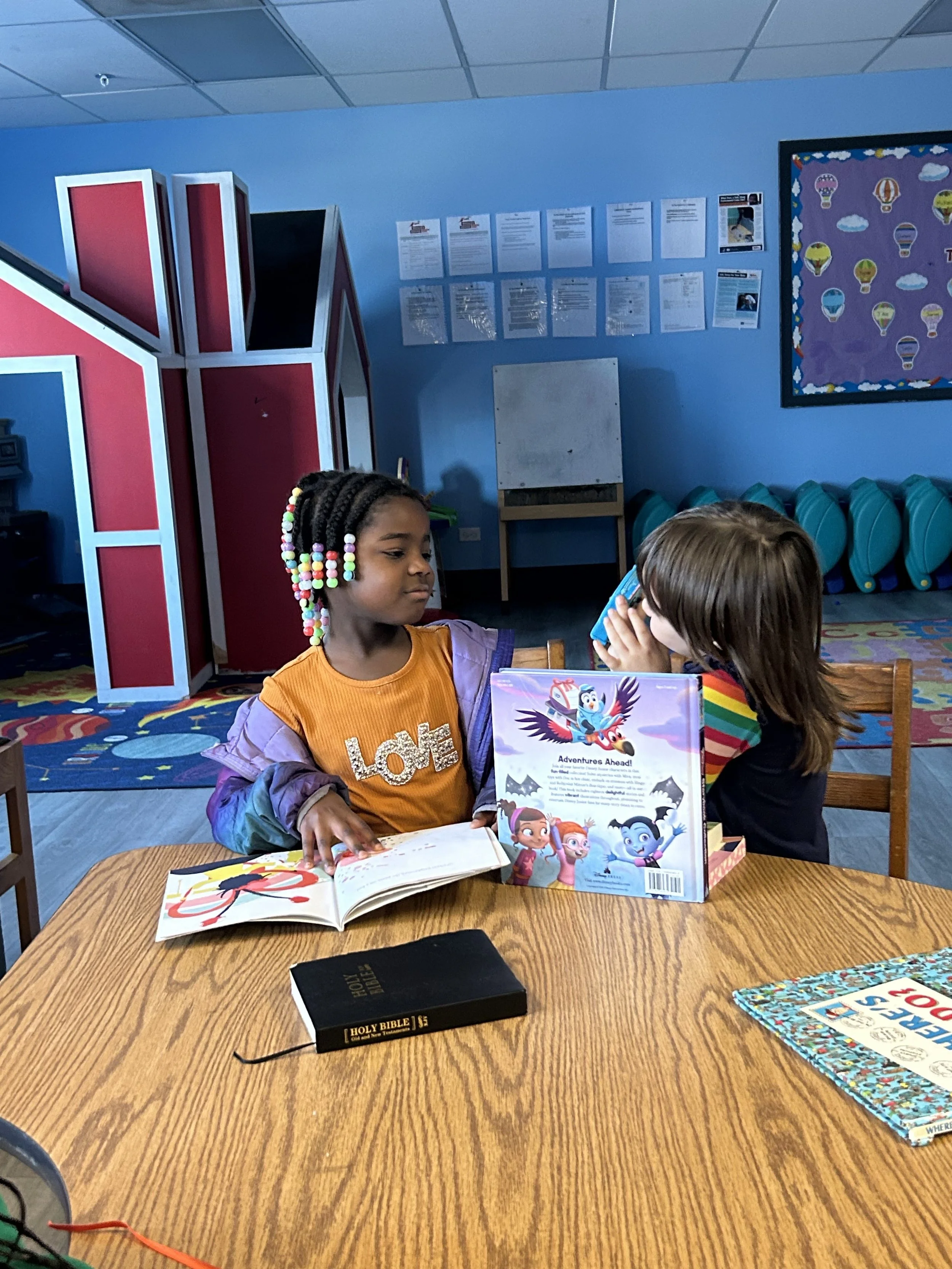 Two young girls sit at a wooden table reading books in a colorful classroom. One girl with braided hair and colorful beads wears an orange shirt with the word 'Love' and is looking at a book, while the other girl with long brown hair and a rainbow-st