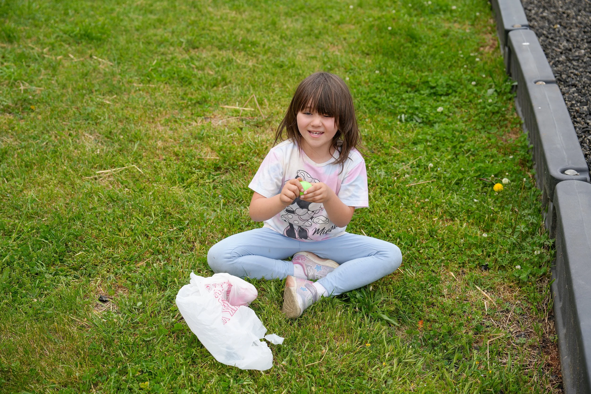 Young girl sitting cross-legged on the grass, holding a green plastic egg, with a crumpled white plastic bag nearby, next to a curb with yellow flowers and small plants.