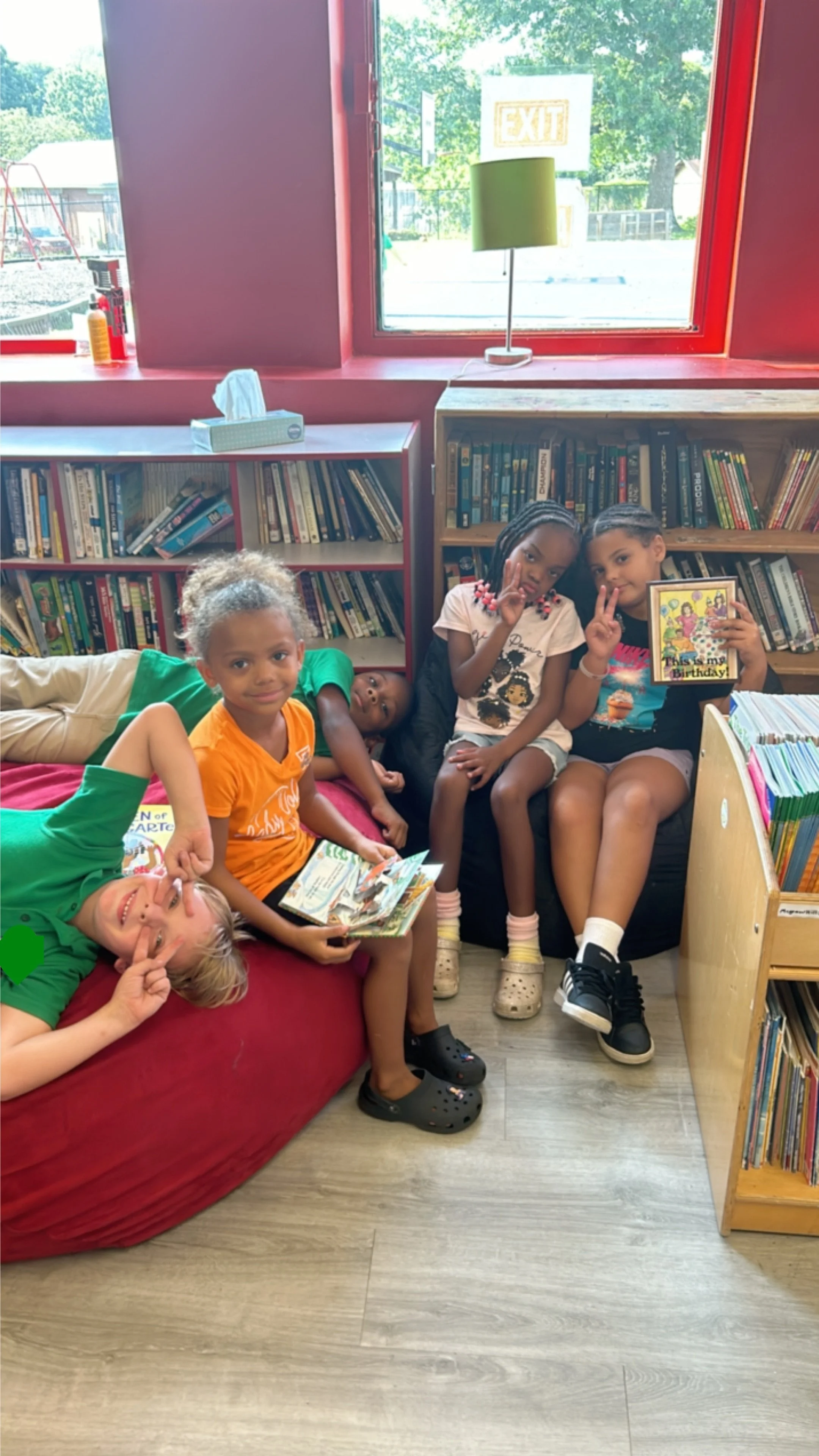 Five children sitting and lying in a library, holding books and making peace signs. There are bookshelves filled with books behind them, and a window with trees outside.