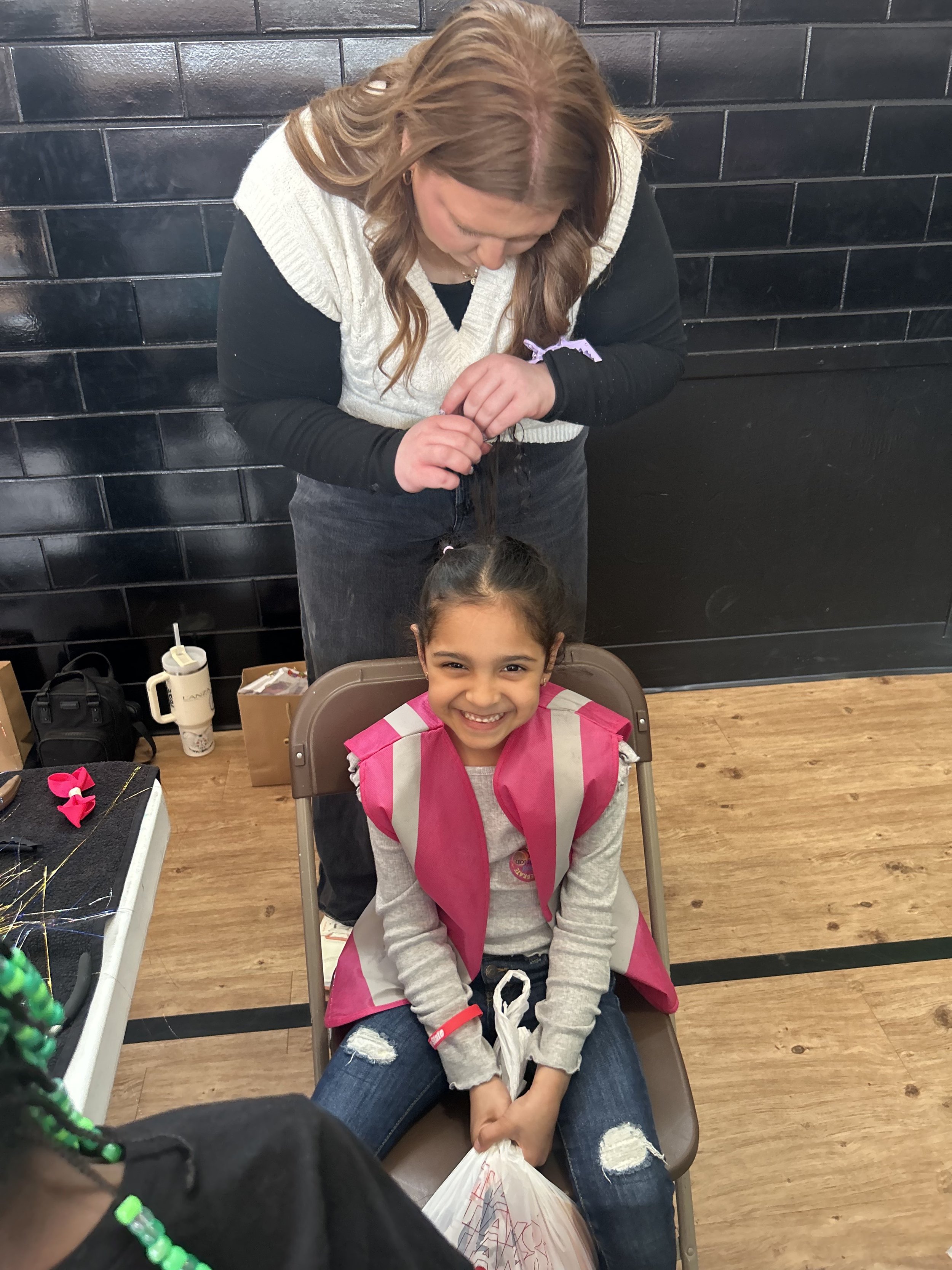 A girl sitting in a chair smiling at the camera while a woman braids her hair.