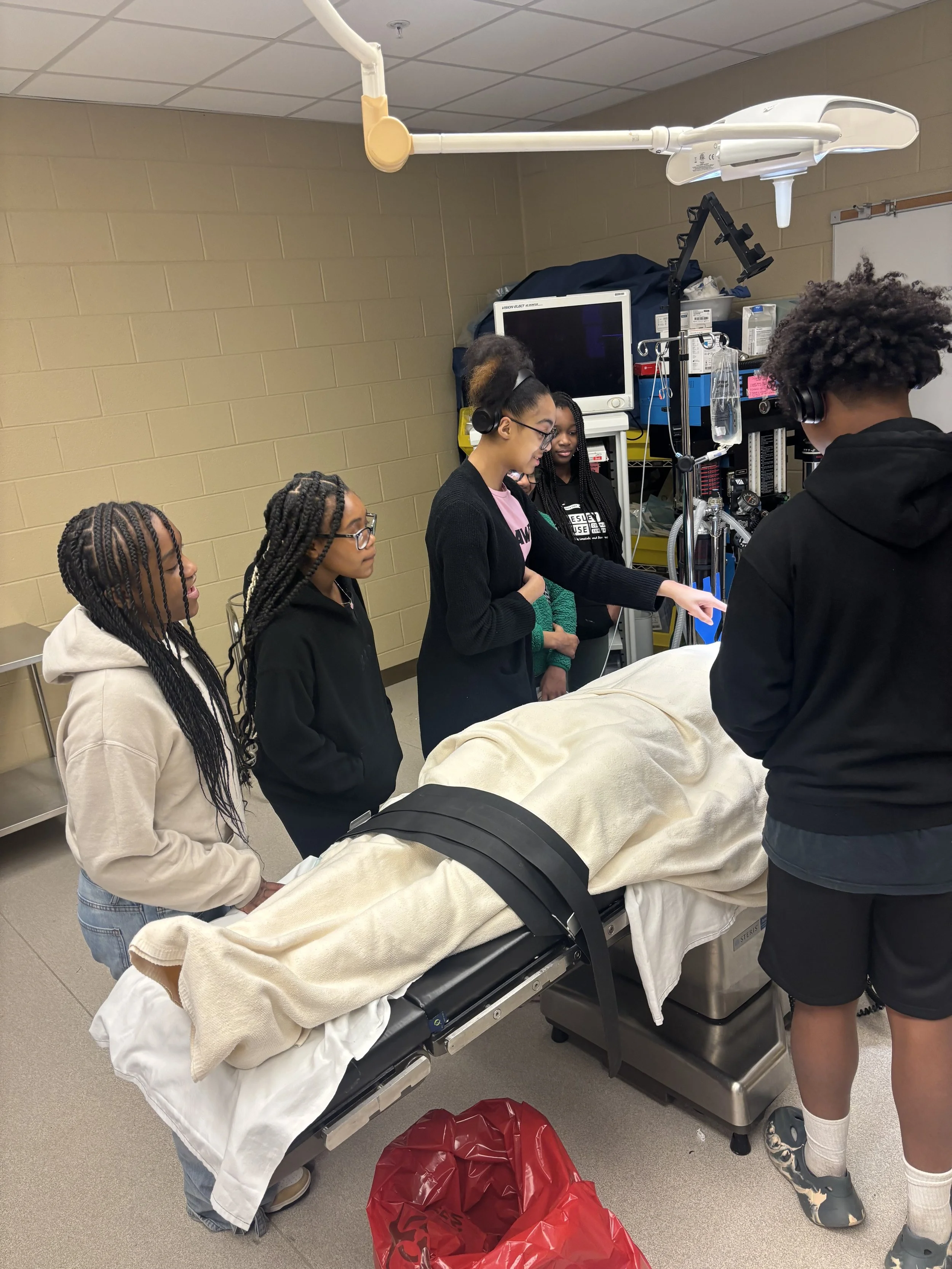 Group of students observing a medical training mannequin in a classroom or hospital room.