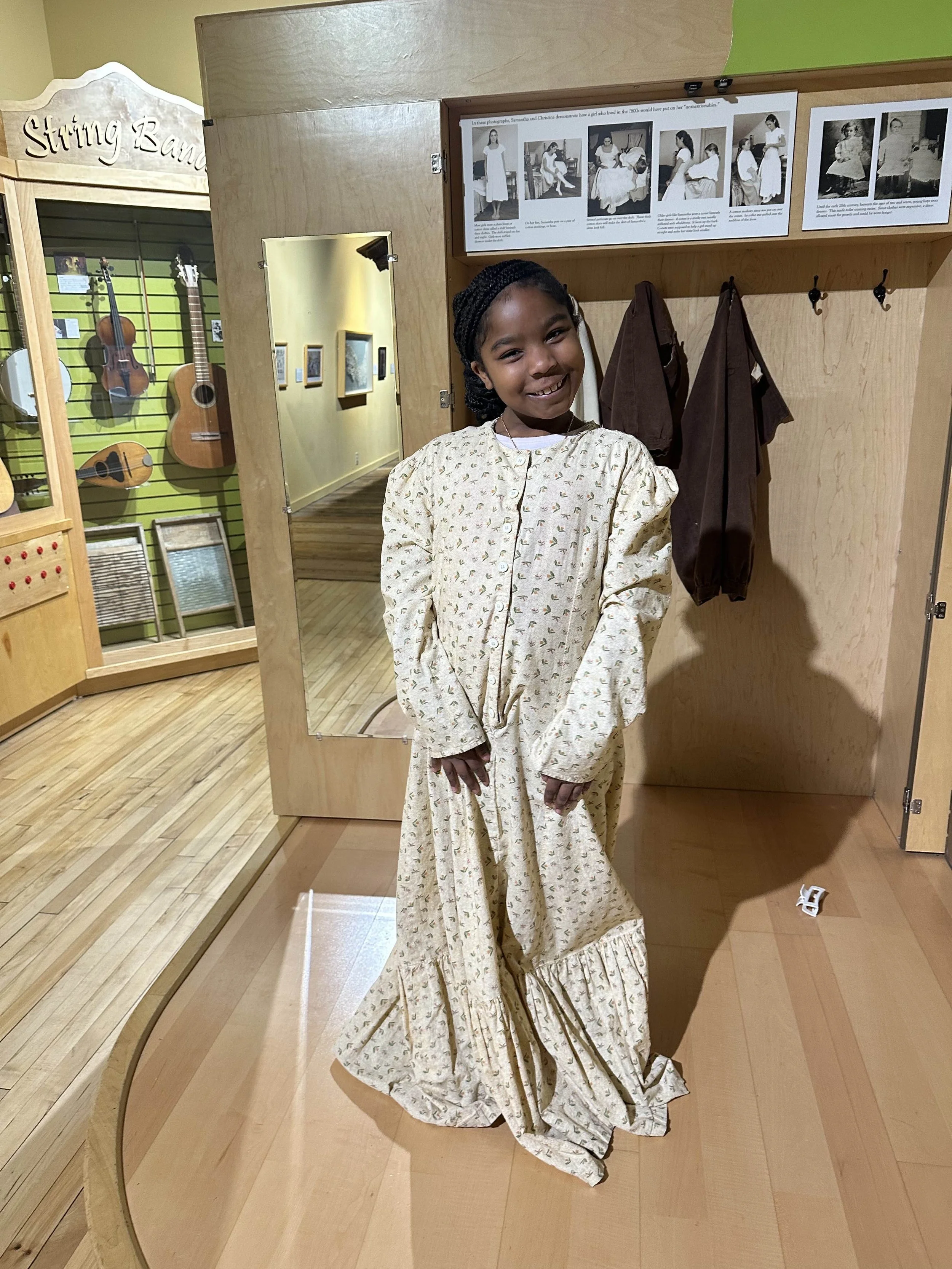 A young girl smiling and standing in a museum exhibit room, wearing a vintage-style cream-colored dress with a floral pattern and ruffled hem, next to a mirror and wooden display area.