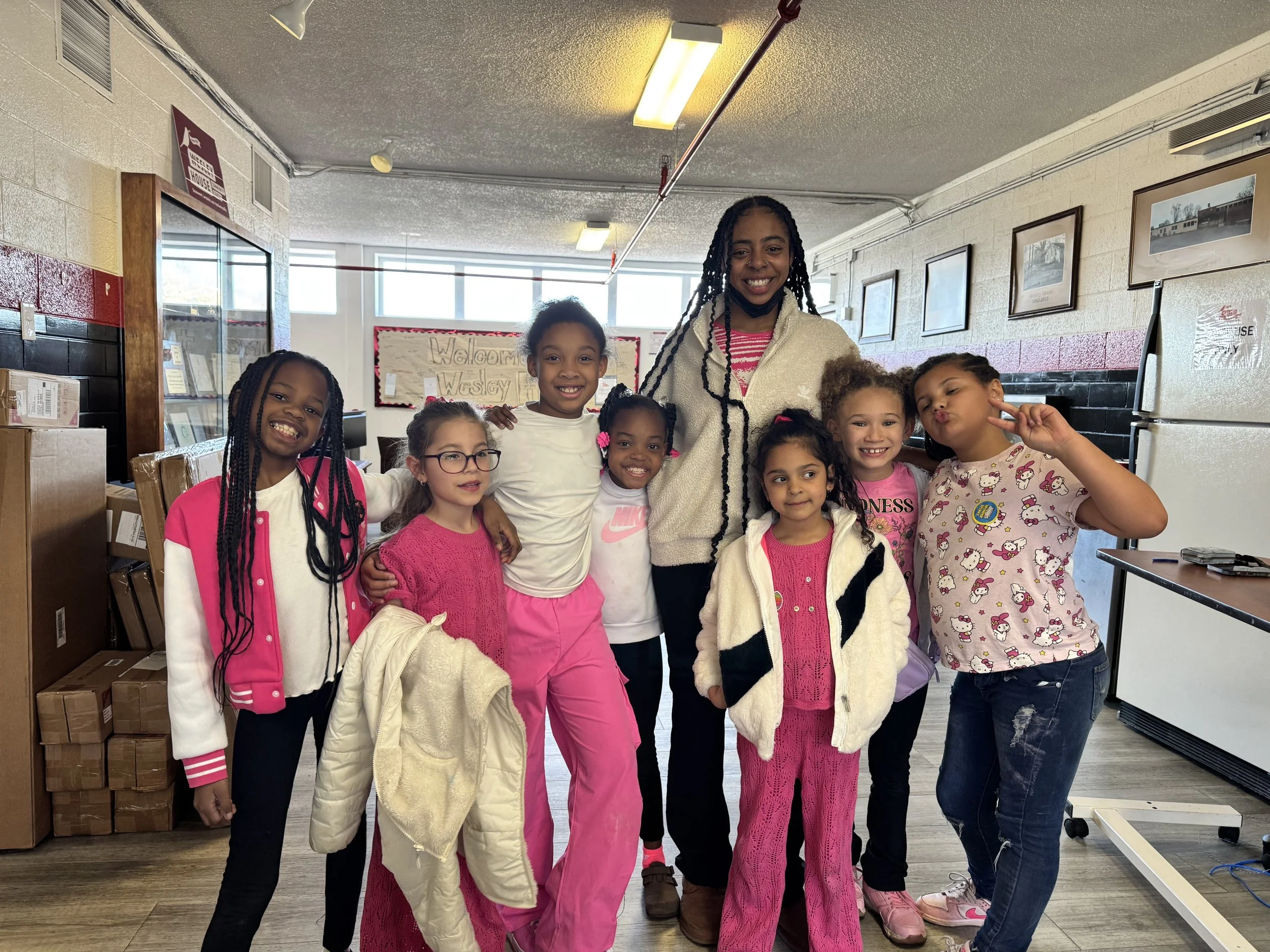 A group of nine smiling young girls and one adult woman posing together indoors in a classroom or community center, with some children making peace signs, wearing casual and colorful clothes, with boxes and framed pictures in the background.