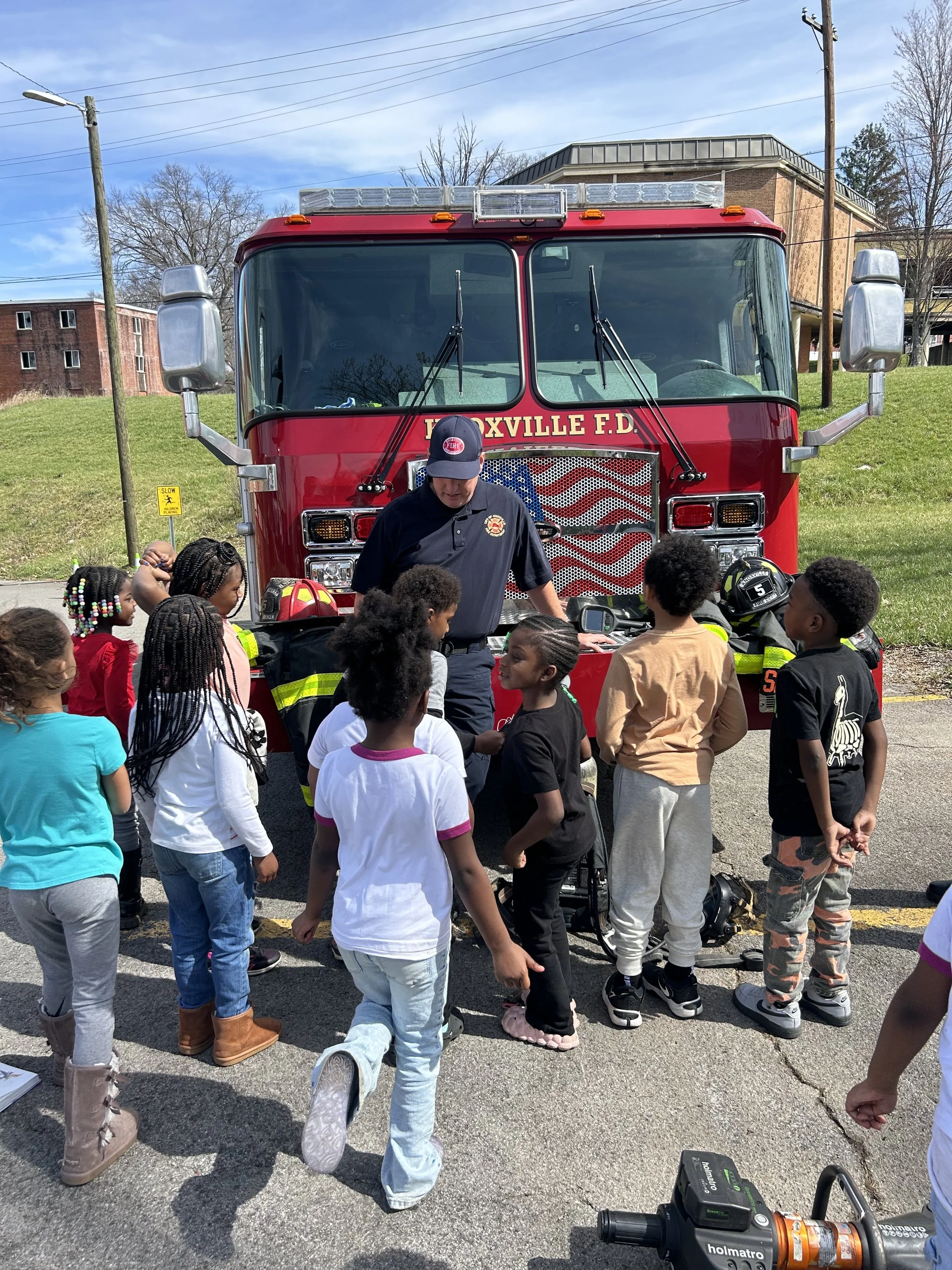 Firefighter demonstrating equipment to a group of children in front of a fire truck.