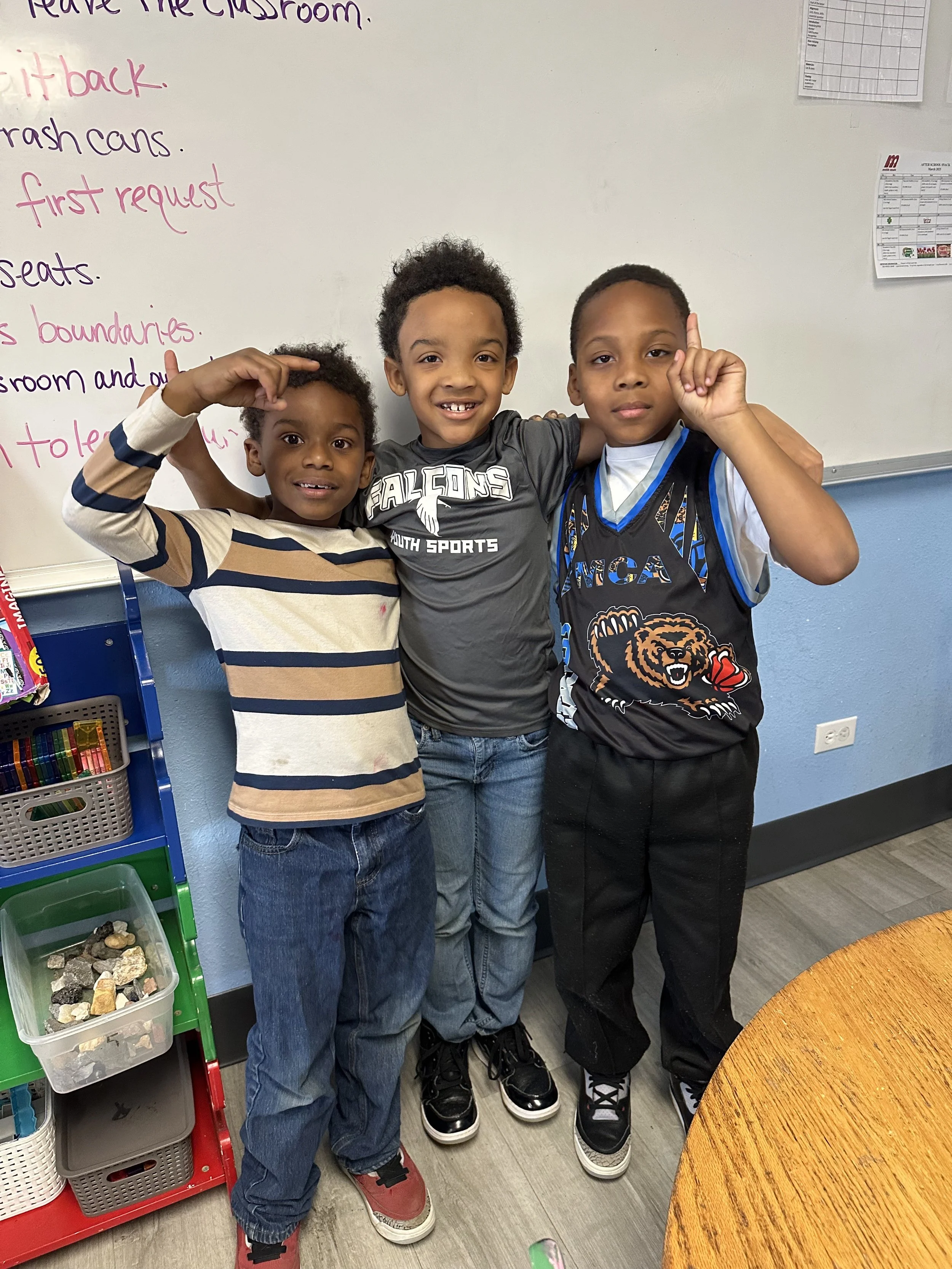 Three young boys standing together in a classroom, smiling at the camera. They are standing in front of a whiteboard with writing on it and a shelving unit with colorful supplies.