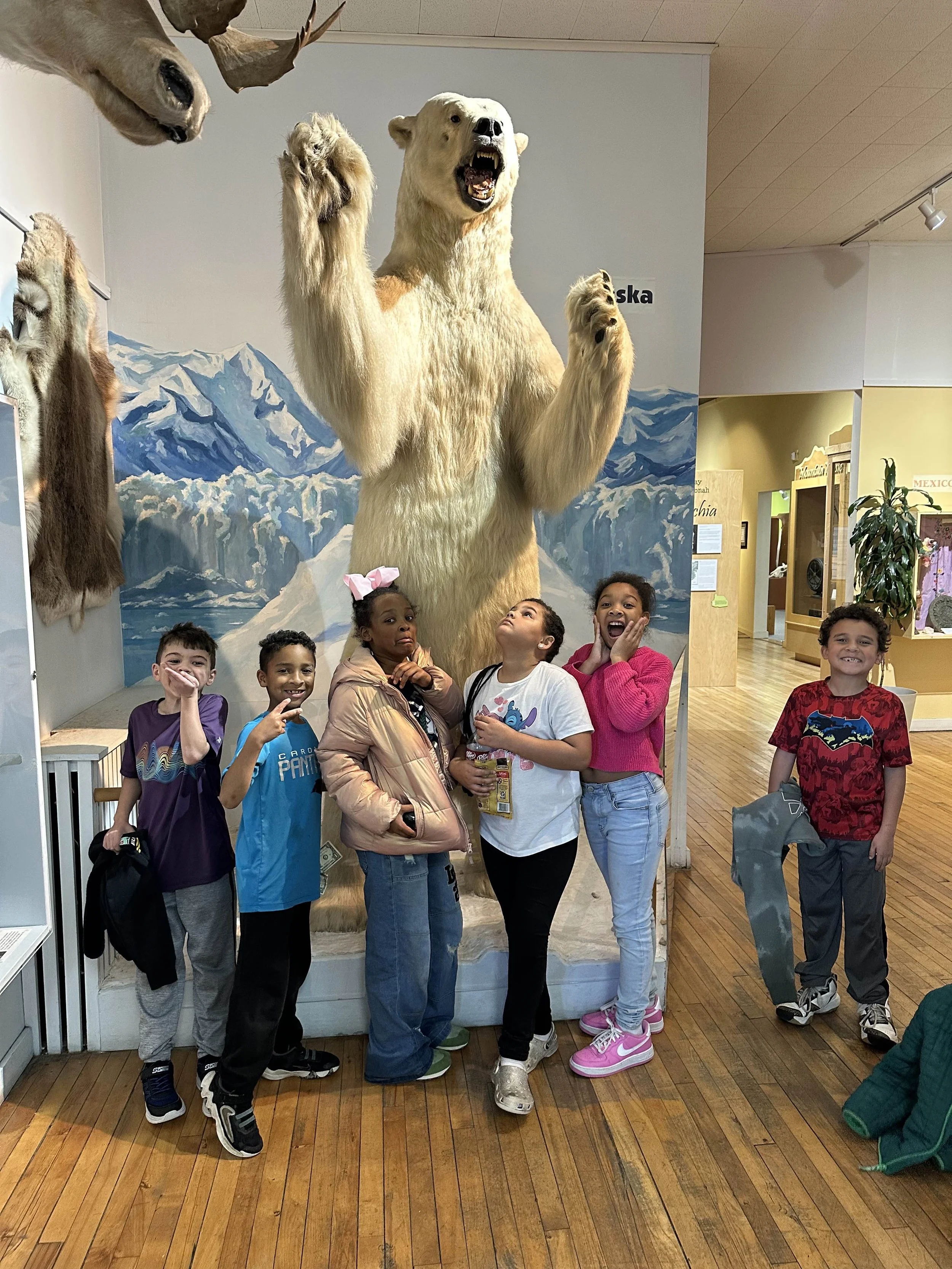 Group of children in a museum posing in front of a large painting of a standing polar bear with claws raised and mouth open, surrounded by other animal taxidermy including a bear and mountain landscape background.