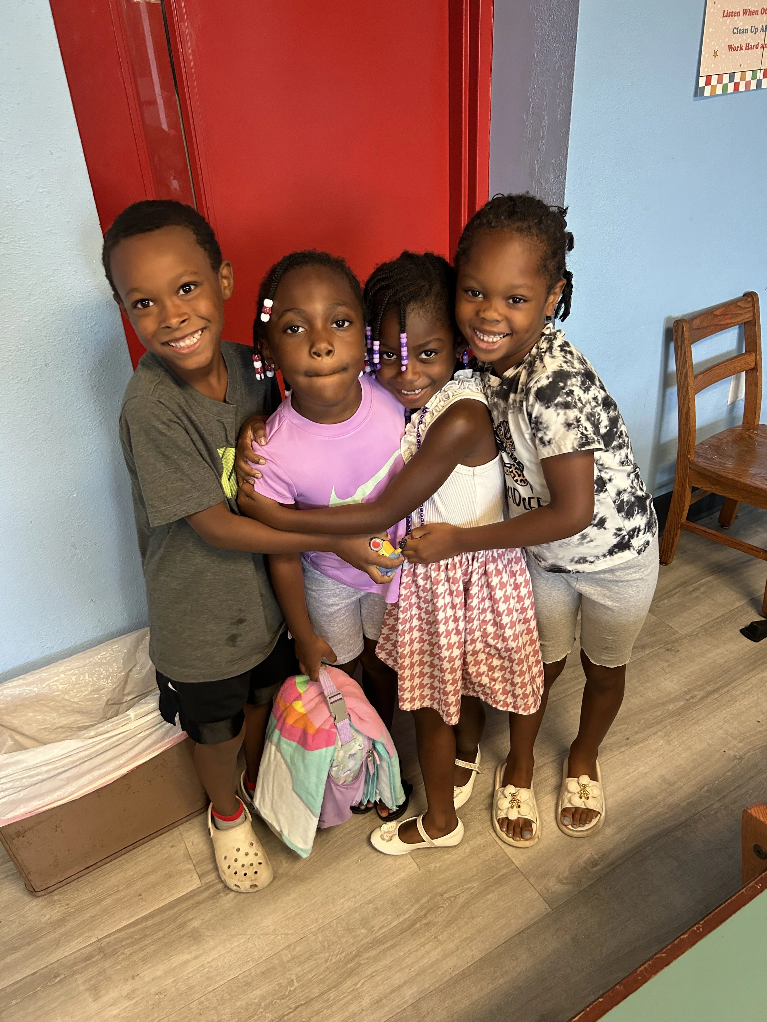 Four children standing close together, smiling and hugging, in an indoor setting near a red door and a blue wall.
