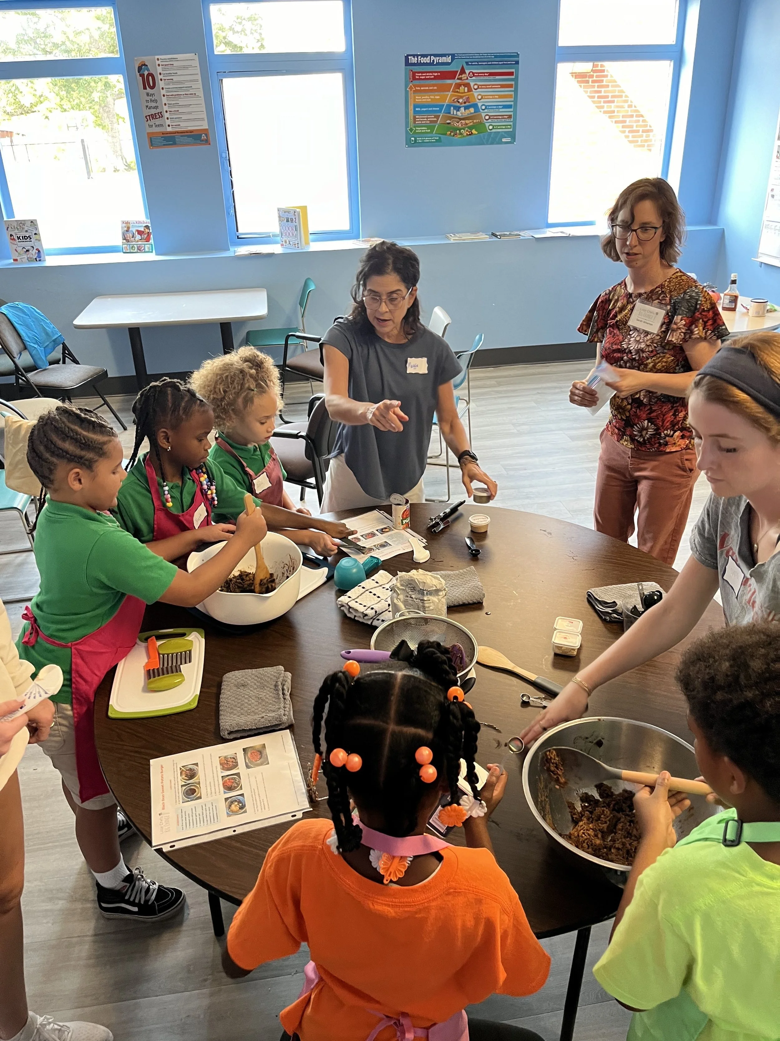 Children participating in a cooking activity at a classroom table, with two adults instructing. The kids are mixing ingredients in bowls, and the room has instructional posters on the walls.