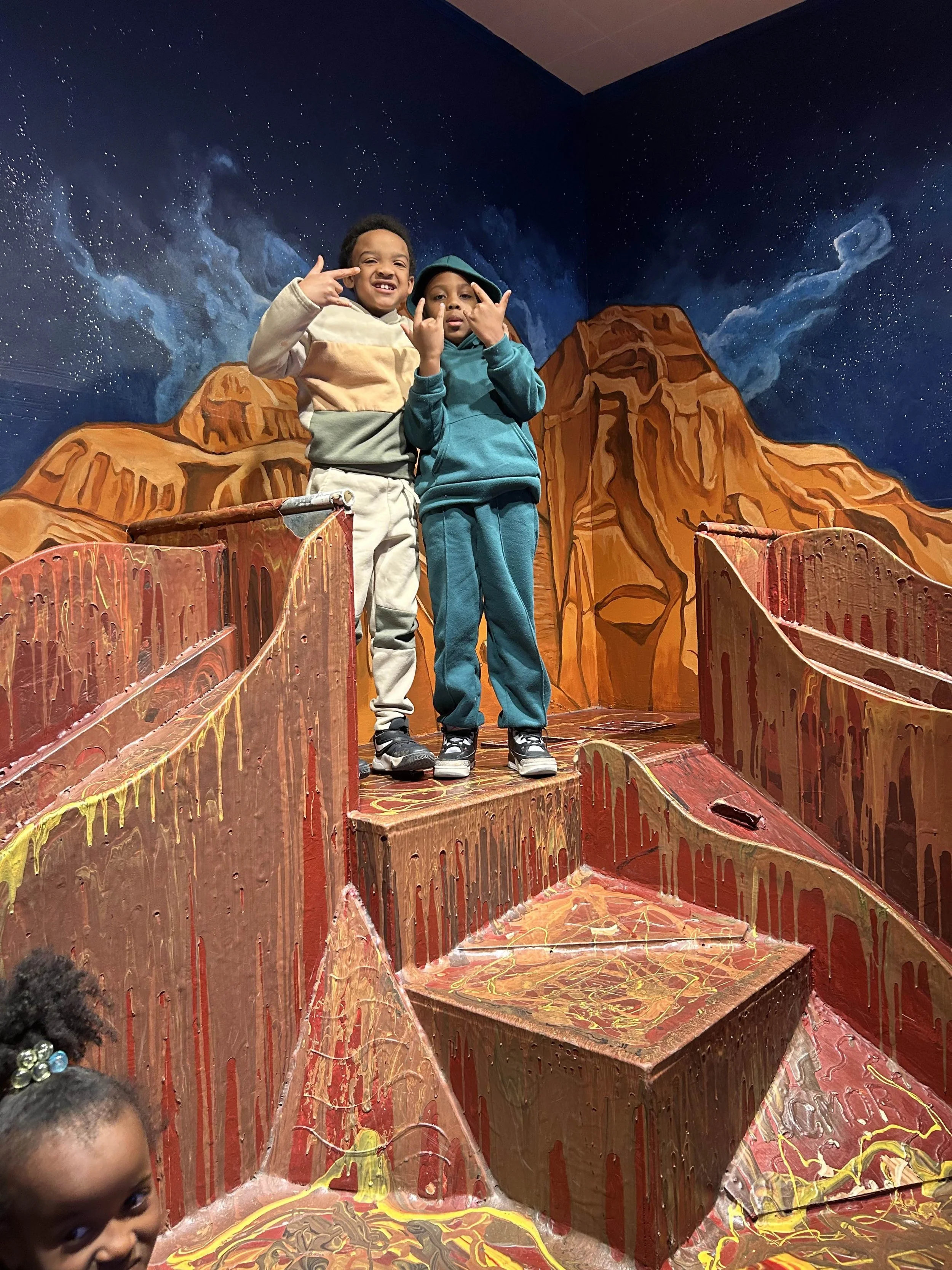 Two young boys standing on a colorful, painted structure resembling a mountain or canyon, with a starry night sky mural in the background.