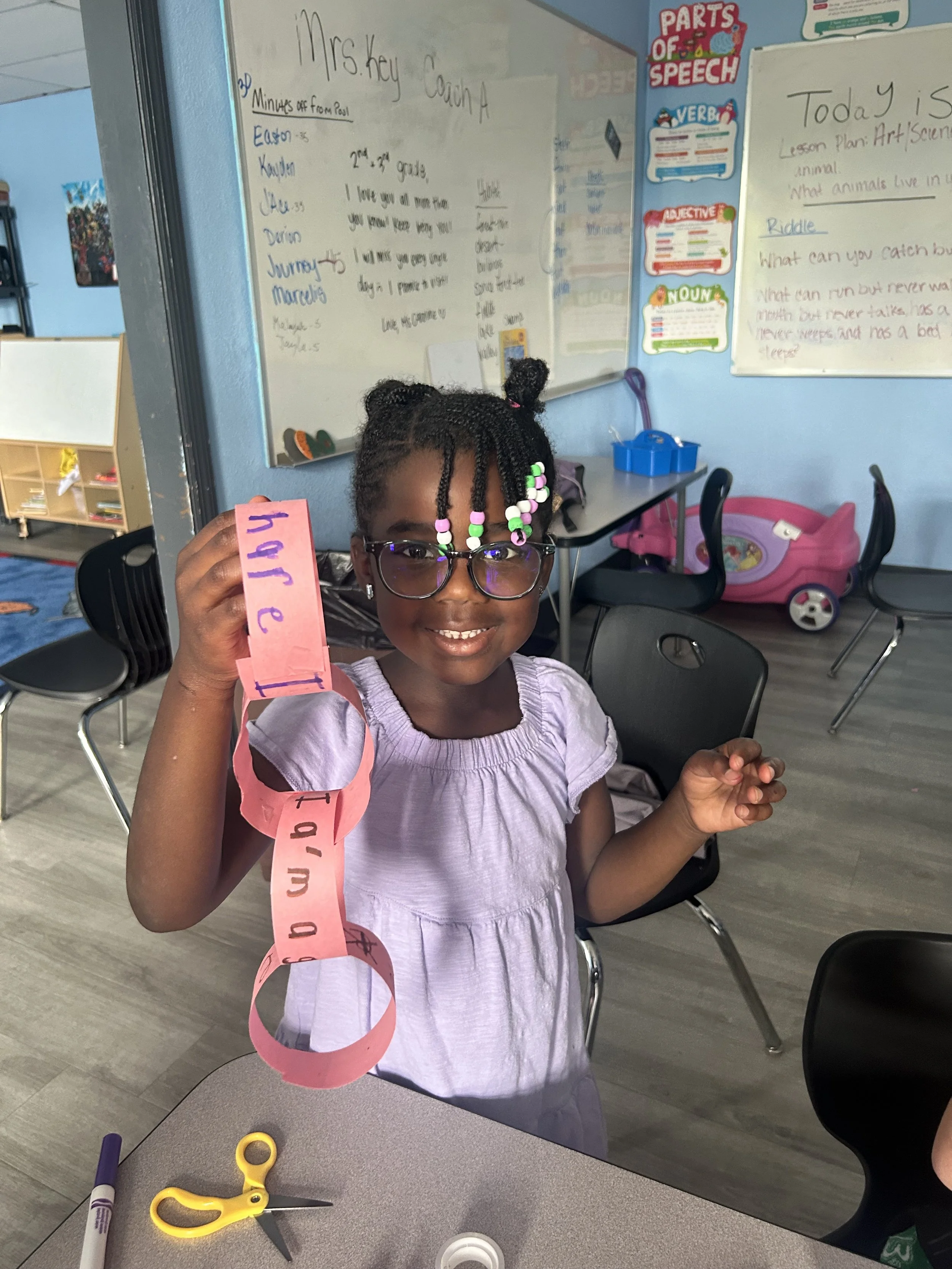 A young girl with glasses and hair decorated with beads is smiling and holding a pink paper chain with cut-out letters. She is seated at a table with scissors and a marker nearby in a classroom, which has colorful posters and a whiteboard.