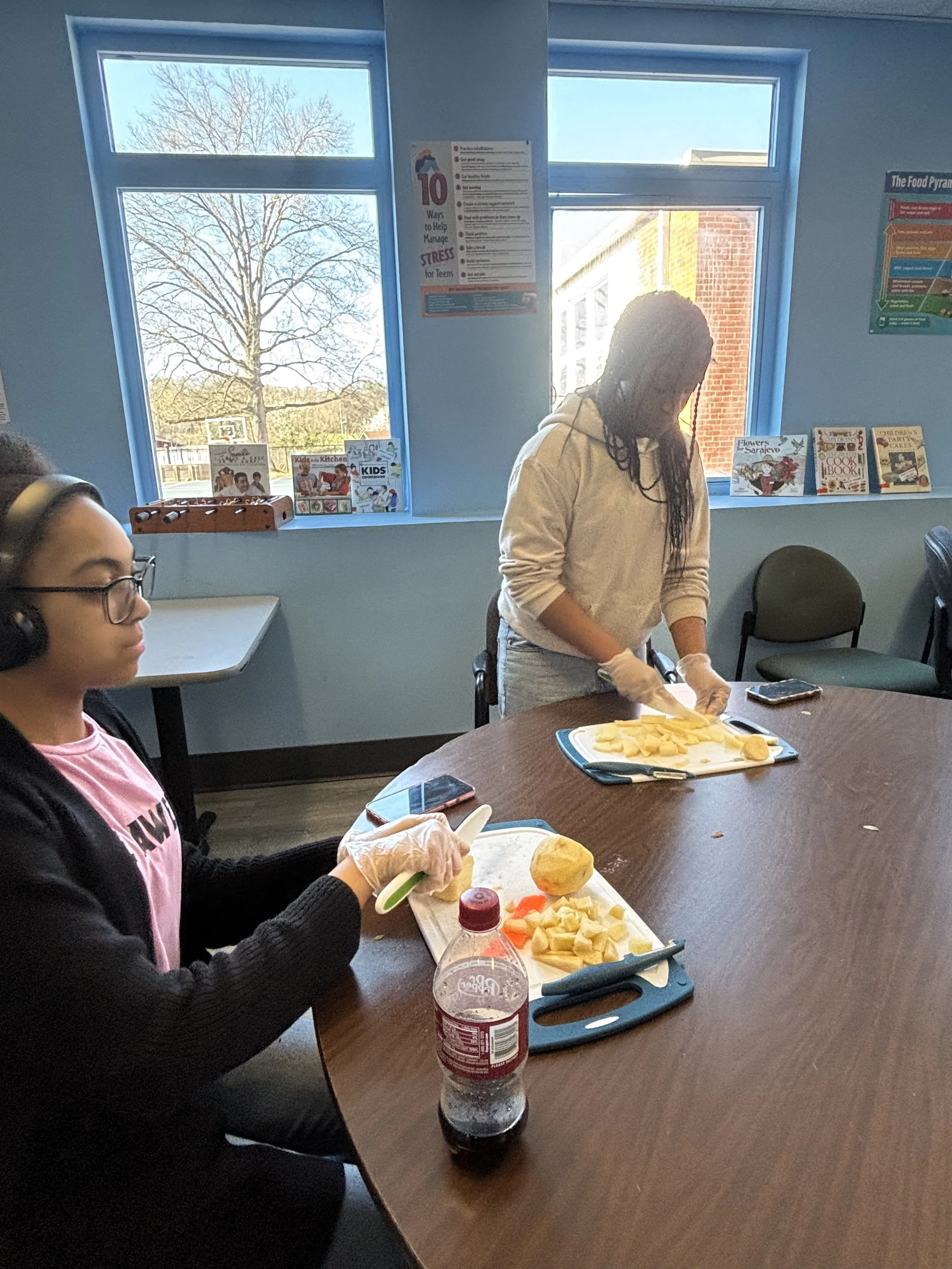 Two women preparing fruit slices at a table in a room with blue walls and windows. One woman is seated wearing glasses and headphones, while the other is standing, slicing fruit.