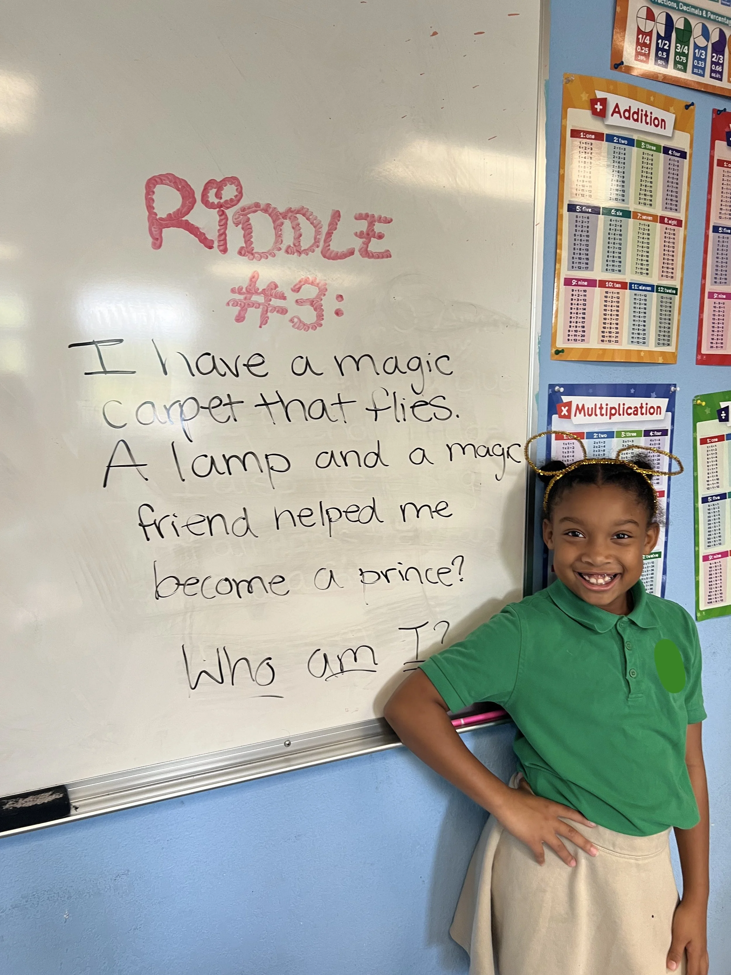 A young girl in a green polo shirt and beige skirt standing beside a whiteboard with a riddle written on it. The whiteboard has colorful charts on the wall behind her.