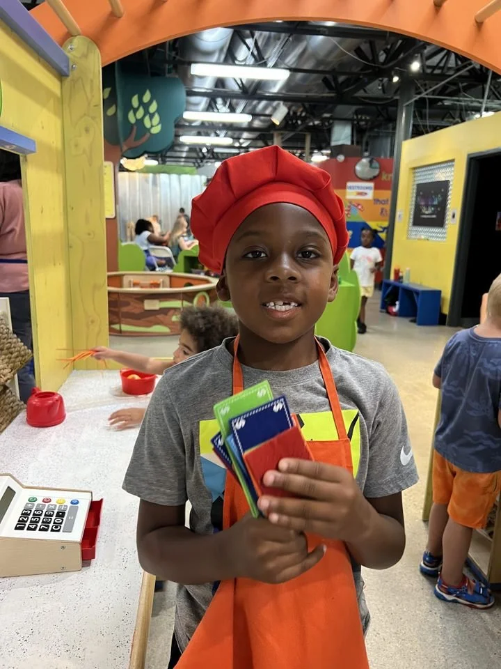 A young boy wearing a red chef hat and gray t-shirt, holding colorful cards, standing inside a children's play area or indoor amusement park.