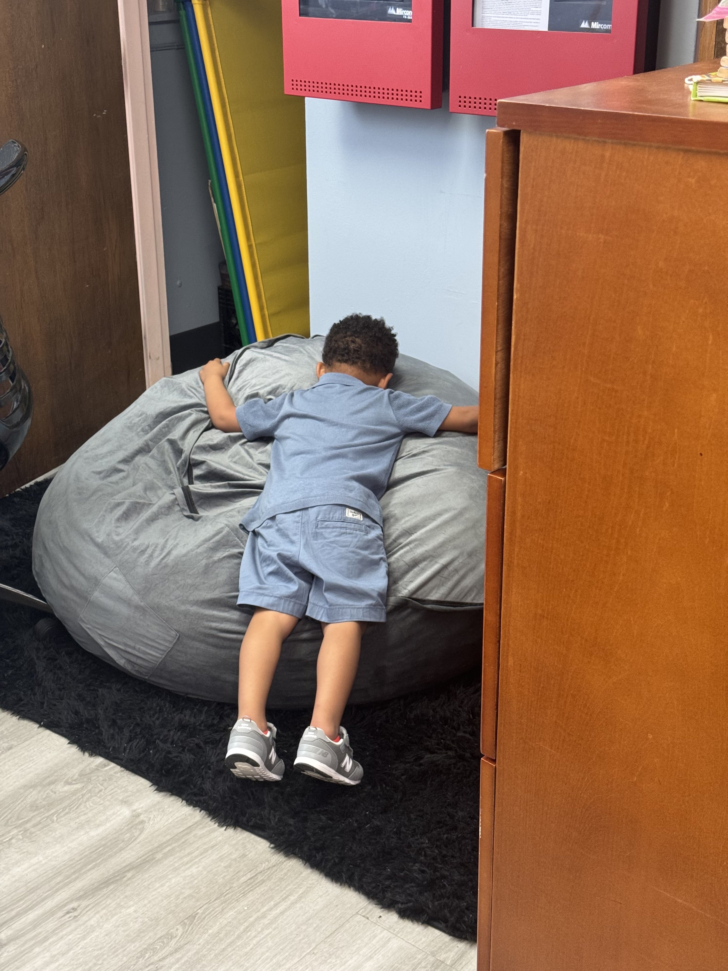 Two young boys are lying face down on a large bean bag chair in a room with various colorful objects, including yellow and blue foam mats leaning against the wall and red storage boxes mounted on the wall.