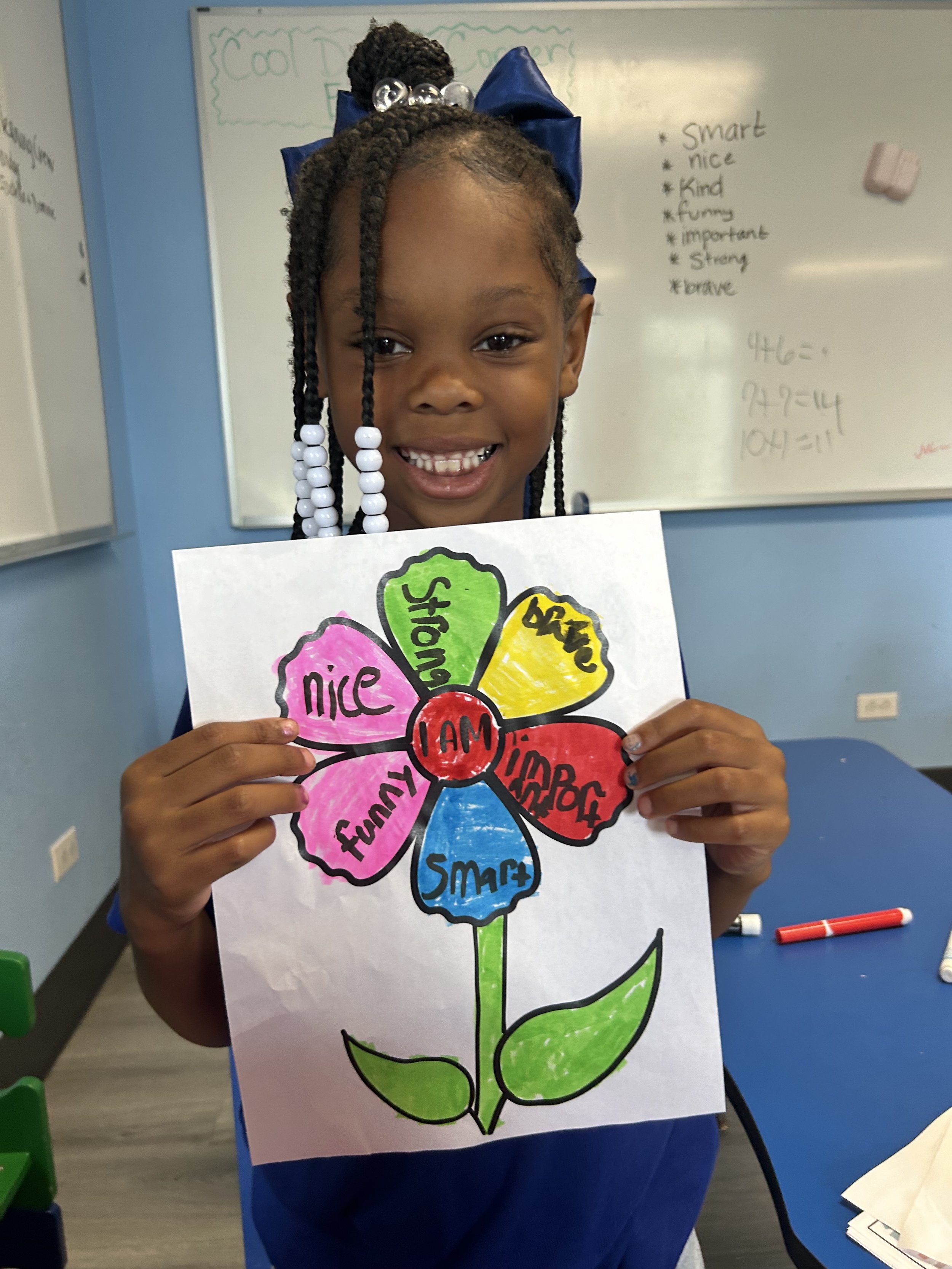 A young girl with braided hair and a blue bow is smiling and holding a colorful drawing of a flower. The flower has seven petals labeled with positive words: 'Smart,' 'Strong,' 'Kind,' 'Funny,' 'Nice,' 'Fun,' and 'Brave,' with a red center that says 