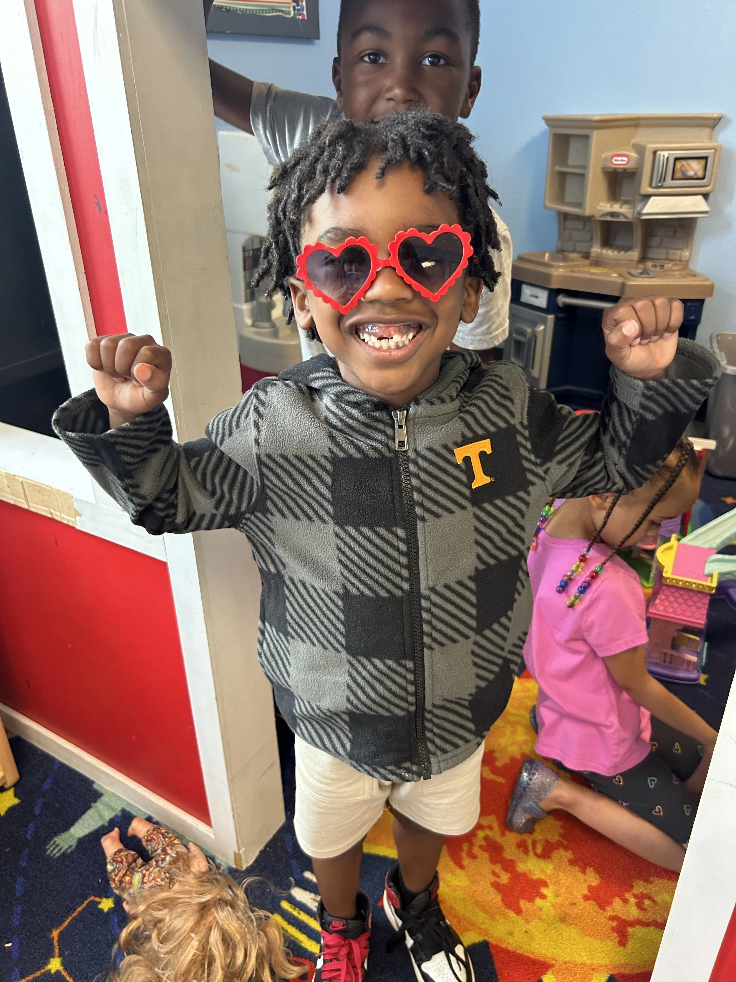 A young boy wearing heart-shaped sunglasses smiling with raised fists inside a colorful playroom, with a boy standing behind him and a girl kneeling nearby.