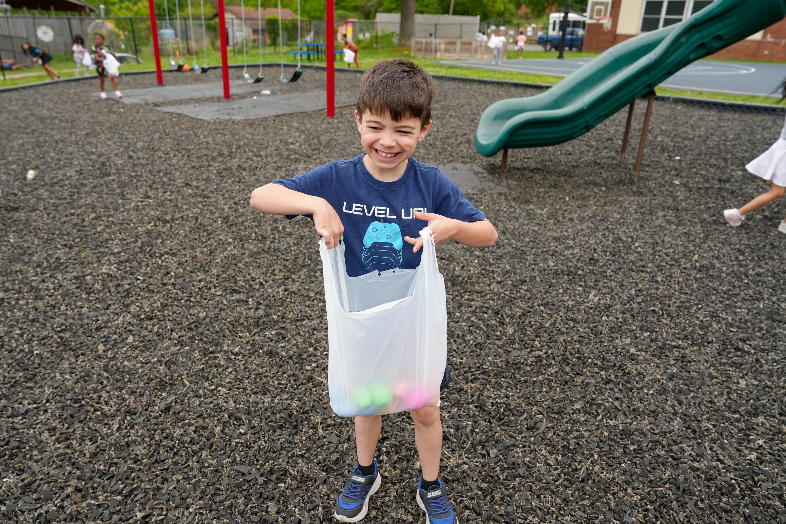 A smiling young boy holding a plastic bag filled with colorful Easter eggs at a playground. In the background, there are children playing, a swing set, a slide, and a basketball court.