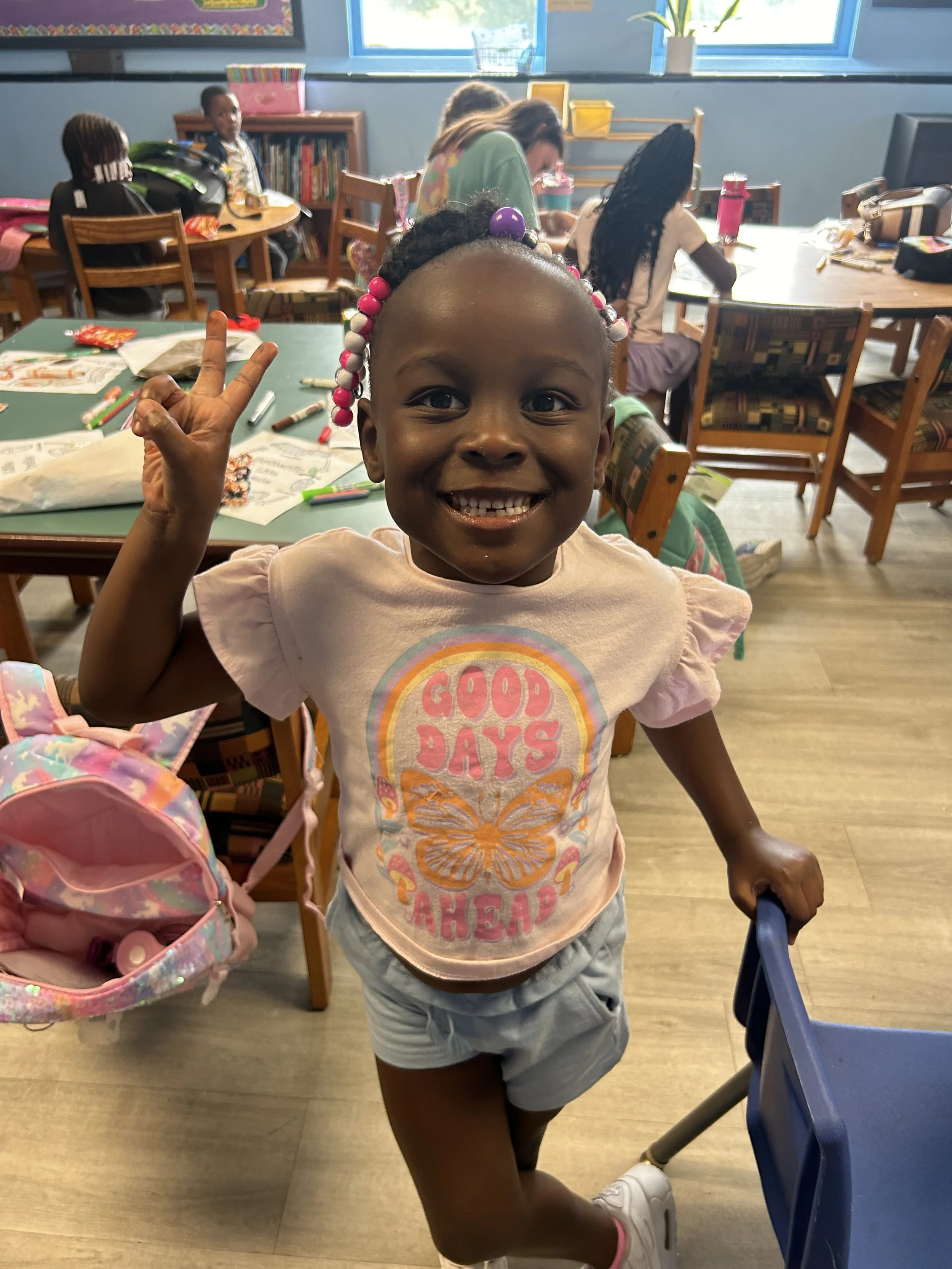 A young girl smiling and making a peace sign with her right hand, holding a small blue shopping cart with her left hand, in a classroom filled with other children and tables with school supplies.