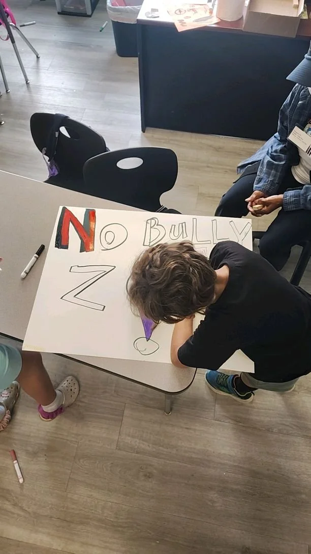 Child writing on a large poster that says 'NO BULLY Z' with a large letter 'N' in red and black, and the other words in black. The child is leaning over the table with a purple marker. Someone's legs and shoes are also visible around the table.