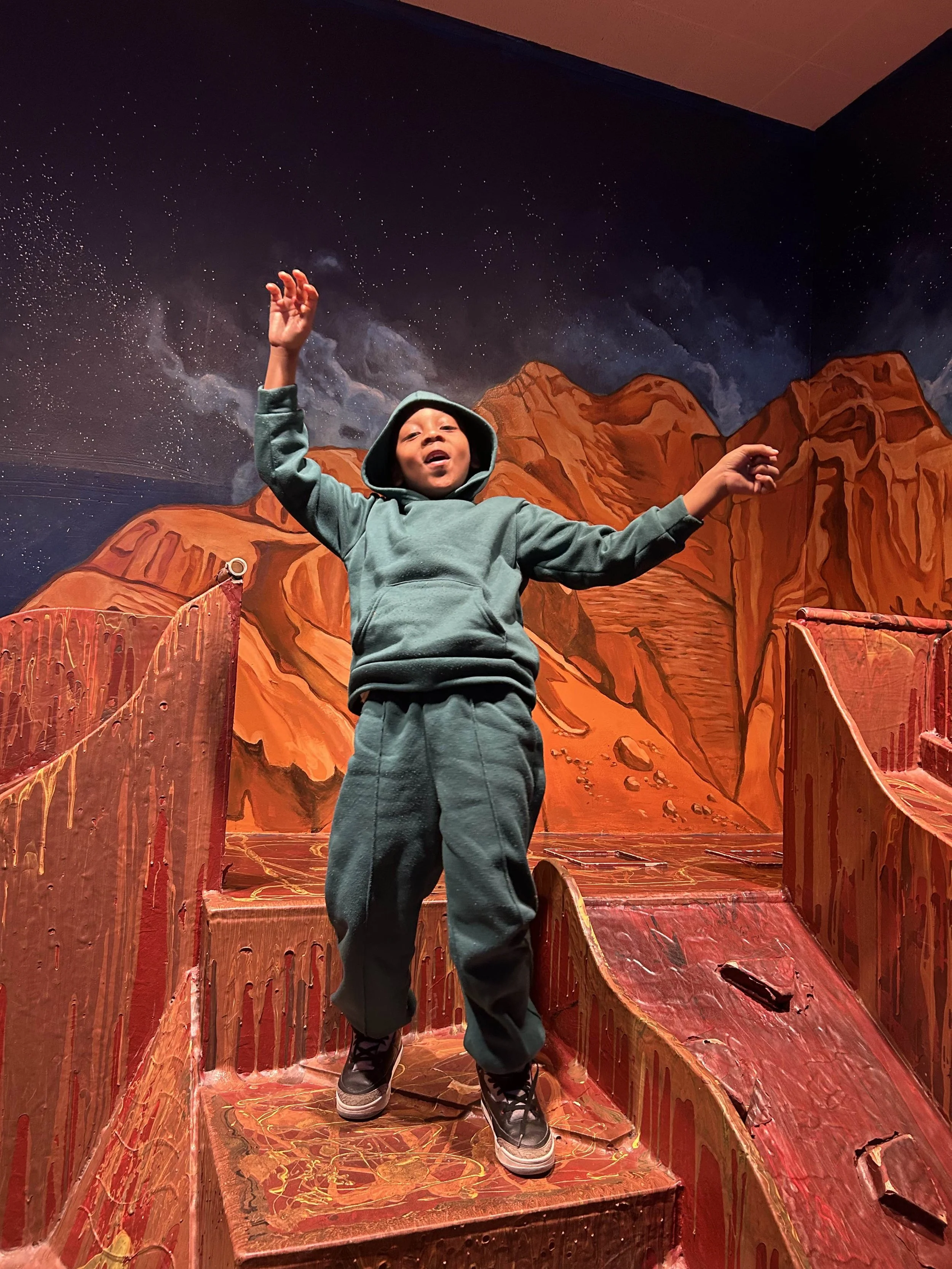 A young boy in green hoodie and pants standing on a colorful painted wooden structure resembling a mountain scene, with a space-themed mural featuring stars and nebulae in the background.