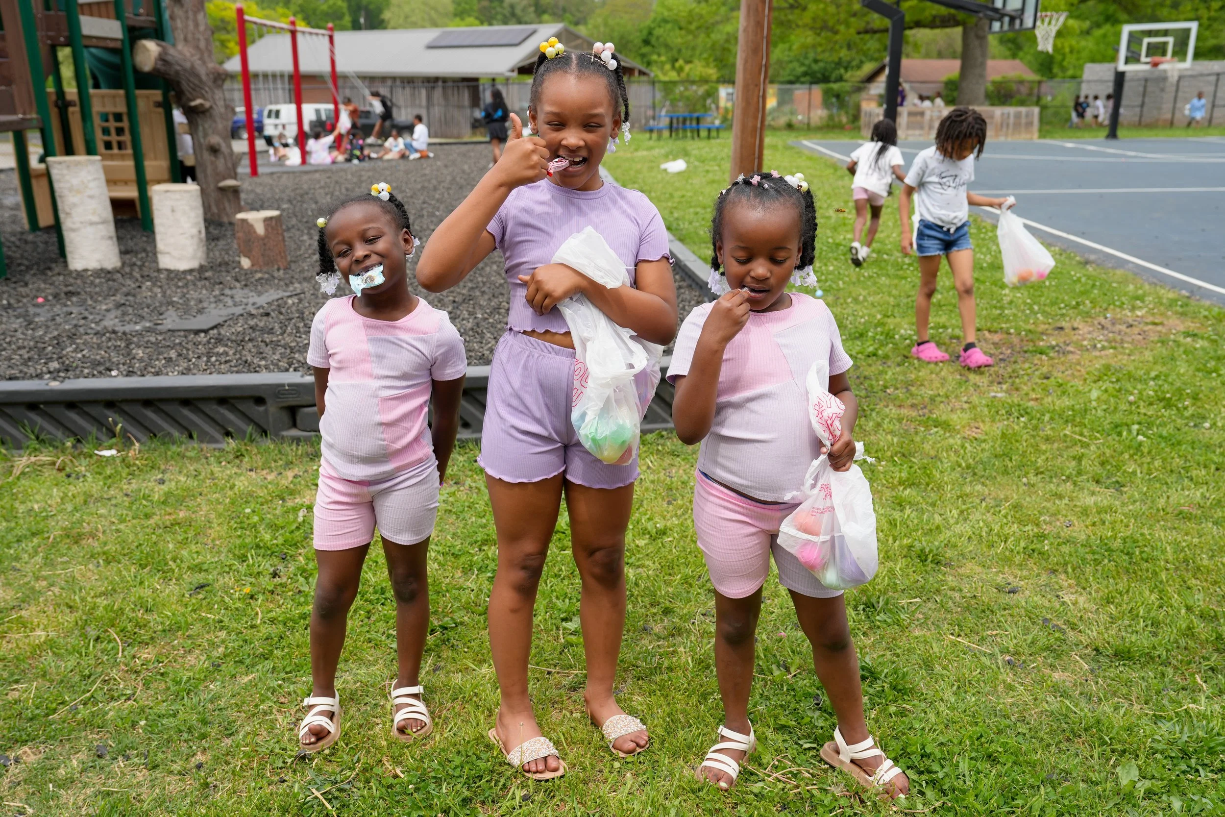 Three young girls enjoying a colorful egg hunt outdoors on a grassy field in a park, with other children playing in the background.