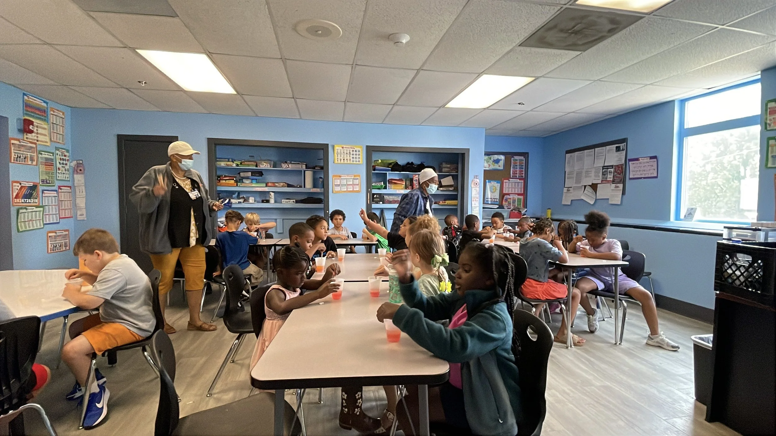 Children in a classroom sitting at tables, drinking juice, and raising their hands, with two teachers walking around, all wearing face masks.