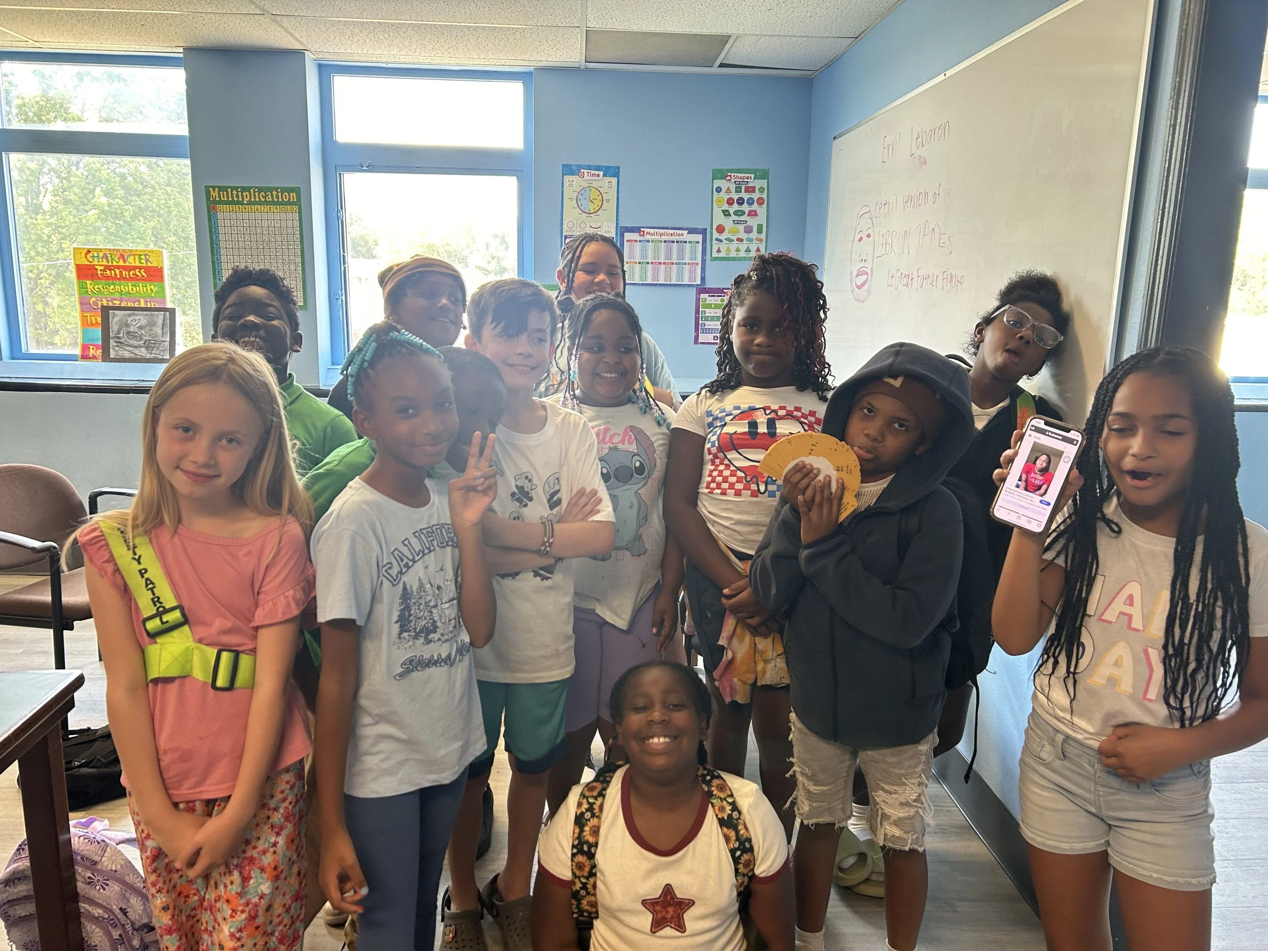 Group of diverse children standing in classroom, some smiling, holding a phone, and making peace signs.