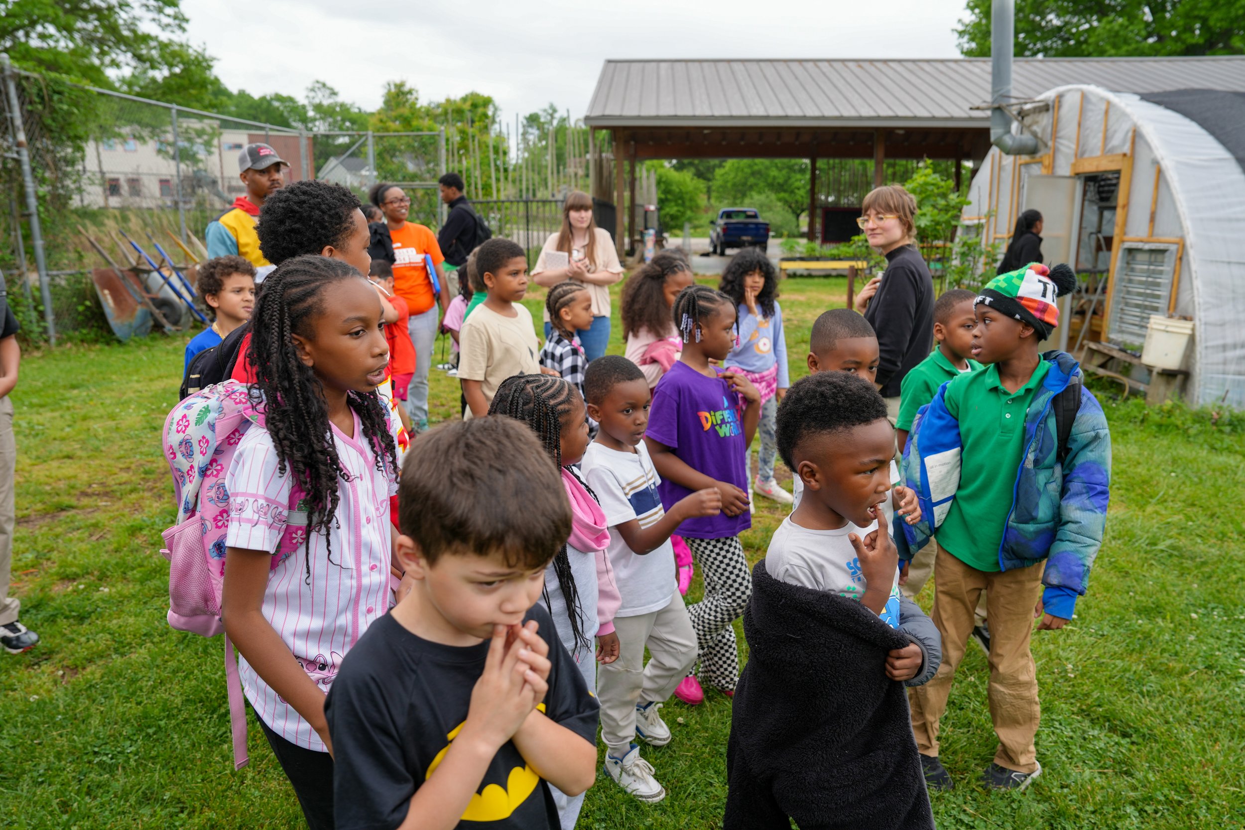 A group of children and a few adults standing outdoors on a grassy area, participating in an educational or recreational activity, with trees and a small building in the background.