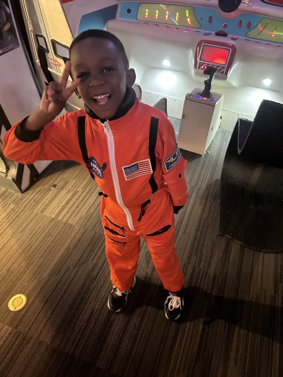 A young boy dressed in an orange astronaut costume standing inside a recreational space-themed arcade game, smiling and making a peace sign with his right hand.
