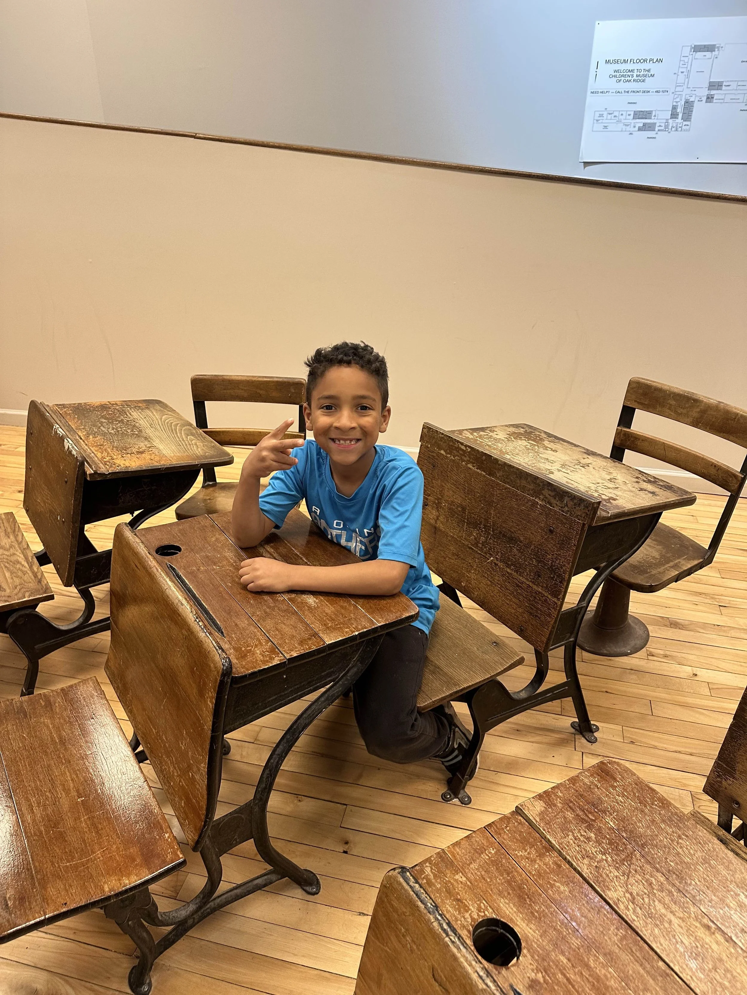 A young boy smiling and making a peace sign while sitting at a vintage wooden school desk in a classroom or museum setting, surrounded by similar desks.