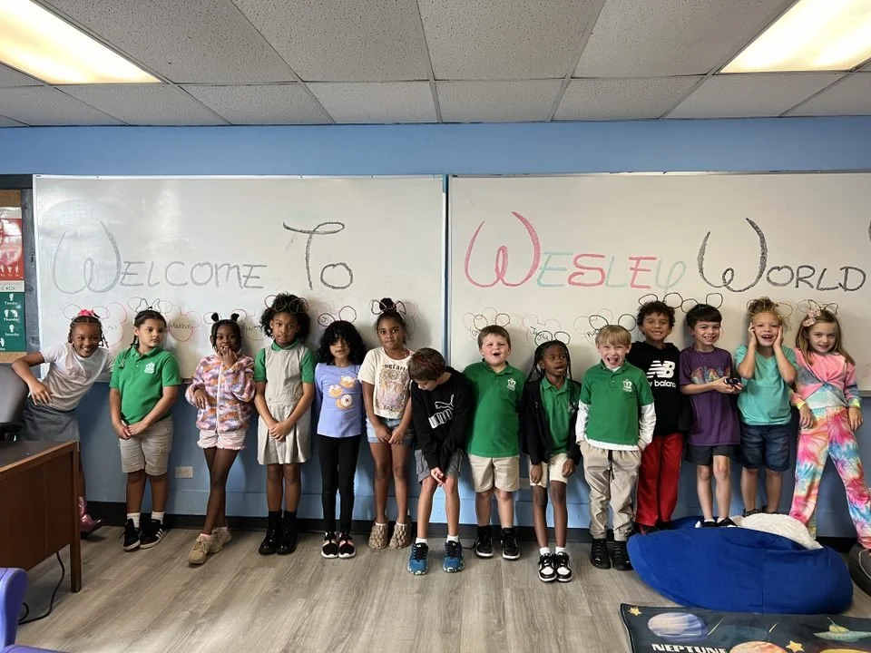 Group of children standing in front of a whiteboard with colorful writing that says 'Welcome to WESLD World' in a classroom.