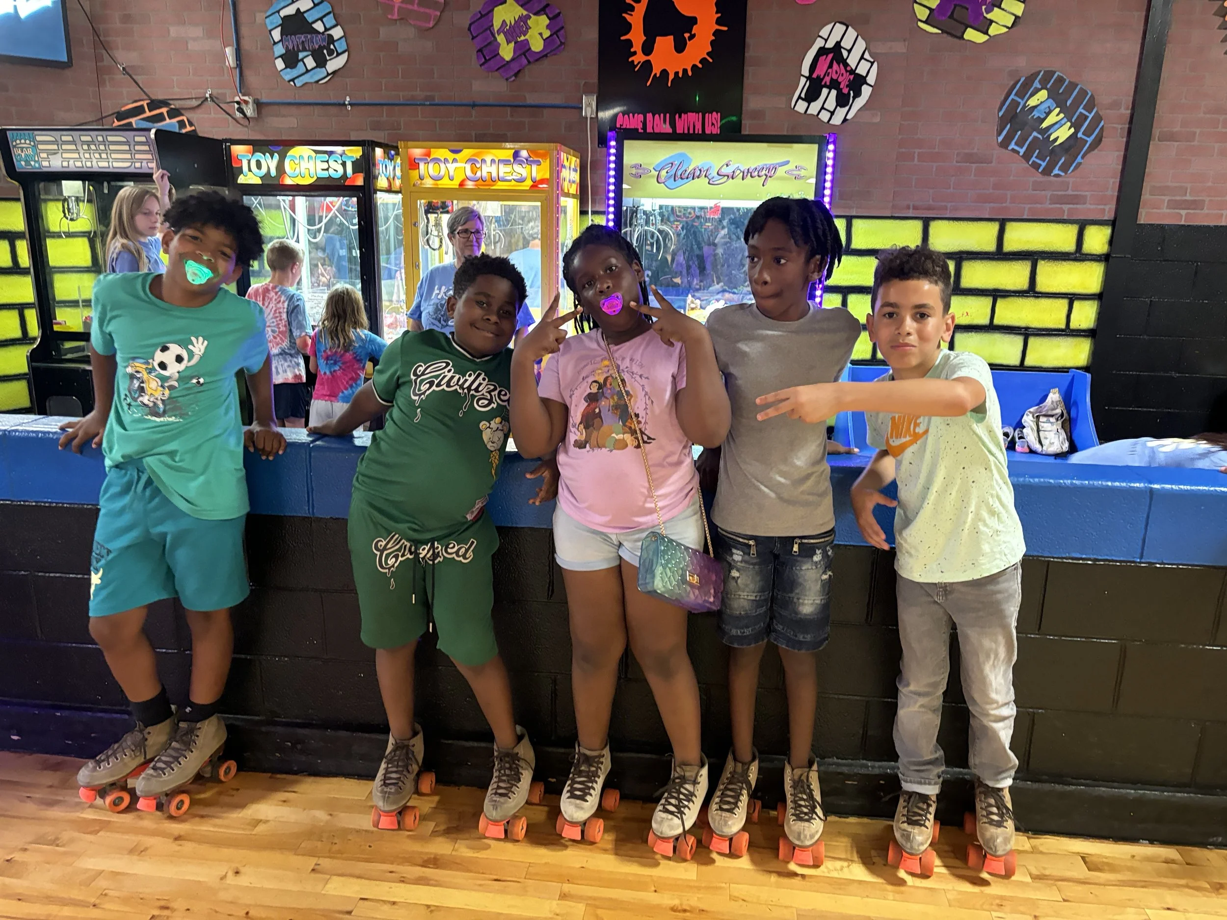 Five kids wearing roller skates lined up at an arcade, with hands on the fence, smiling and posing for the camera. The background features arcade game machines and colorful wall decorations.