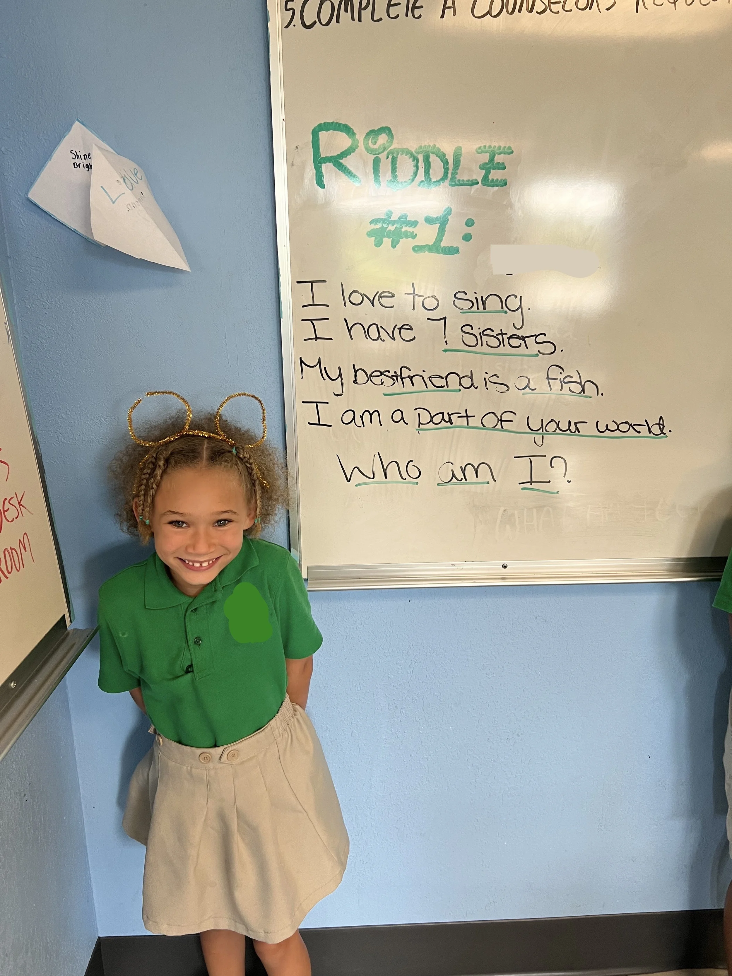 Young girl with curly hair and wearing a green shirt and beige skirt standing in front of a whiteboard with a riddle written on it, smiling at the camera.