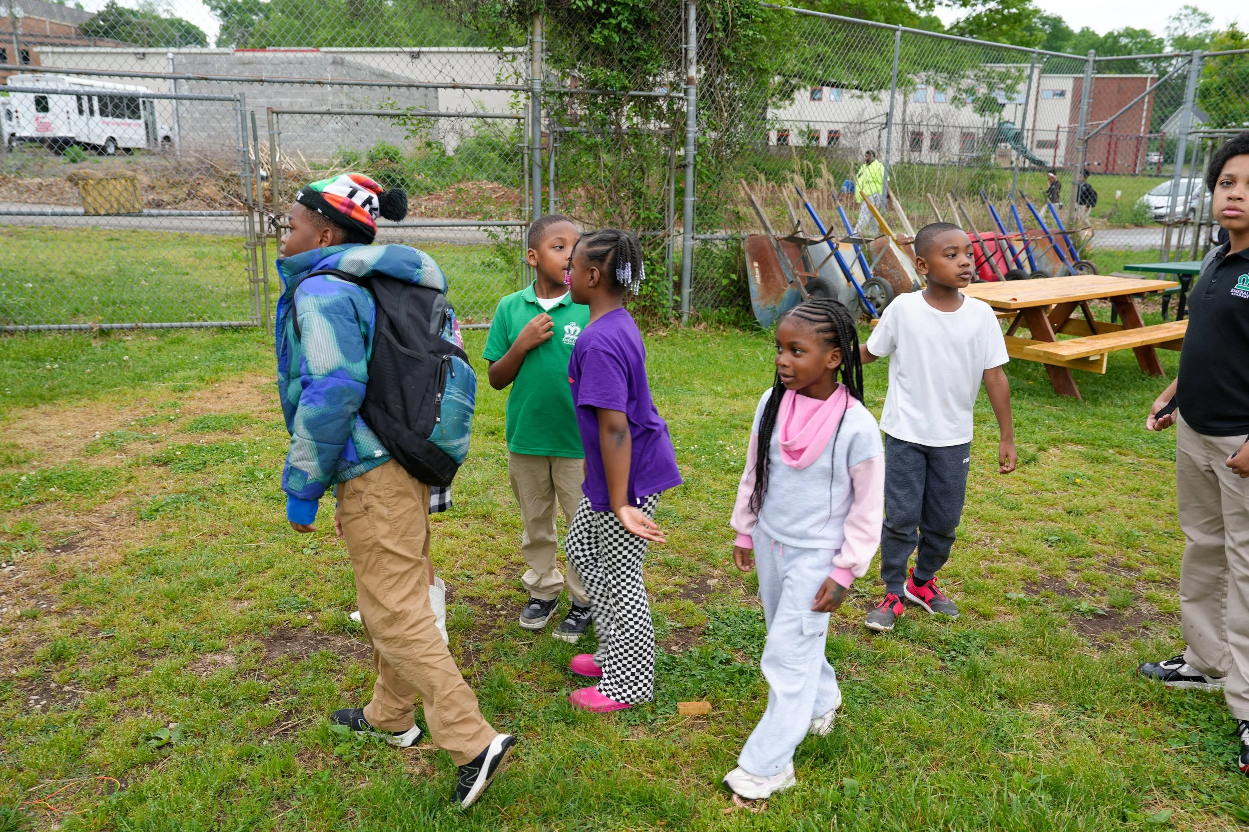 Group of children and an adult standing on grassy area near chain-link fence with playground equipment and wheelbarrows in the background.