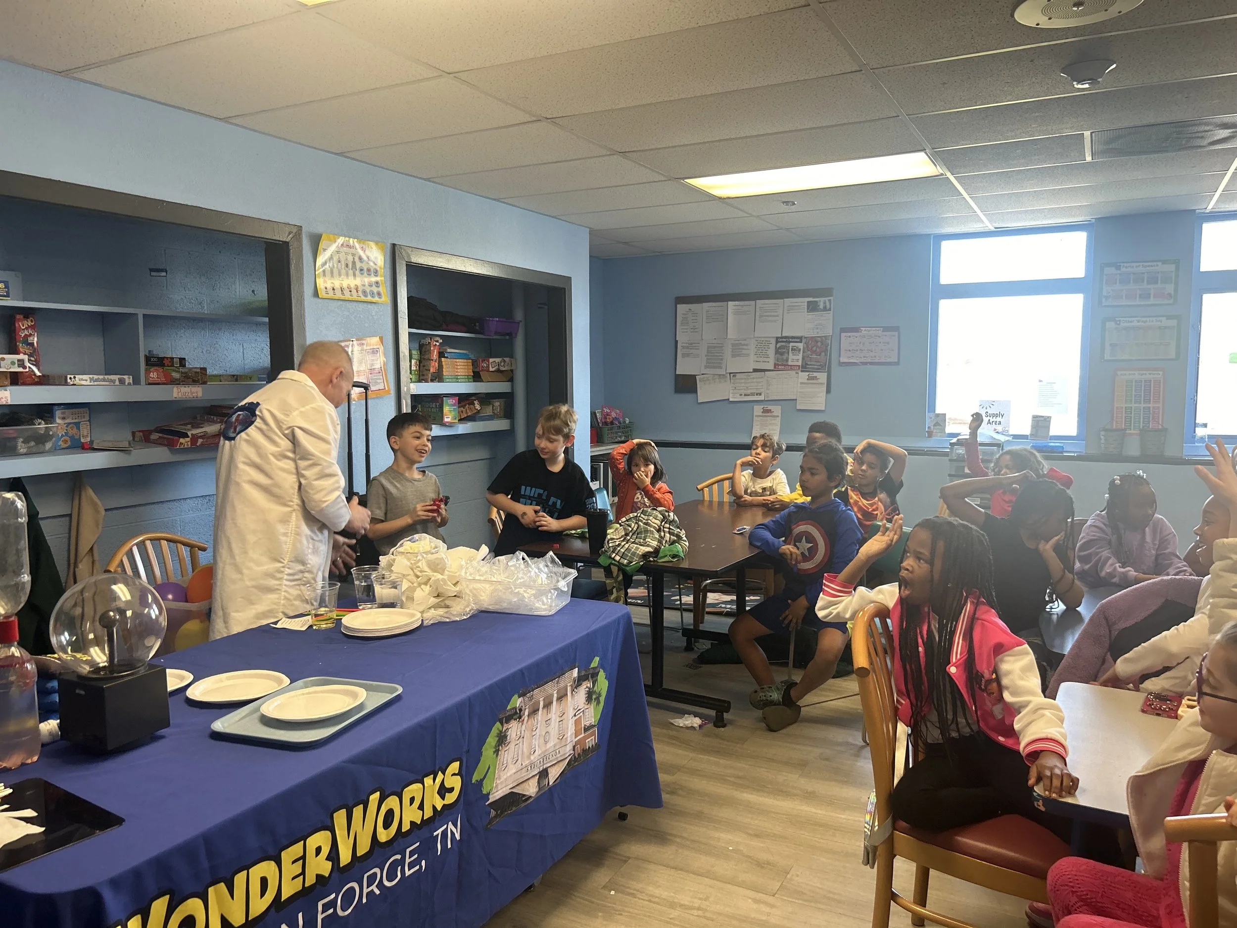 Kids sitting at tables in a classroom, listening to a man in a white lab coat who is conducting a science demonstration with a science kit on a table covered with a blue cloth labeled "WonderWorks."