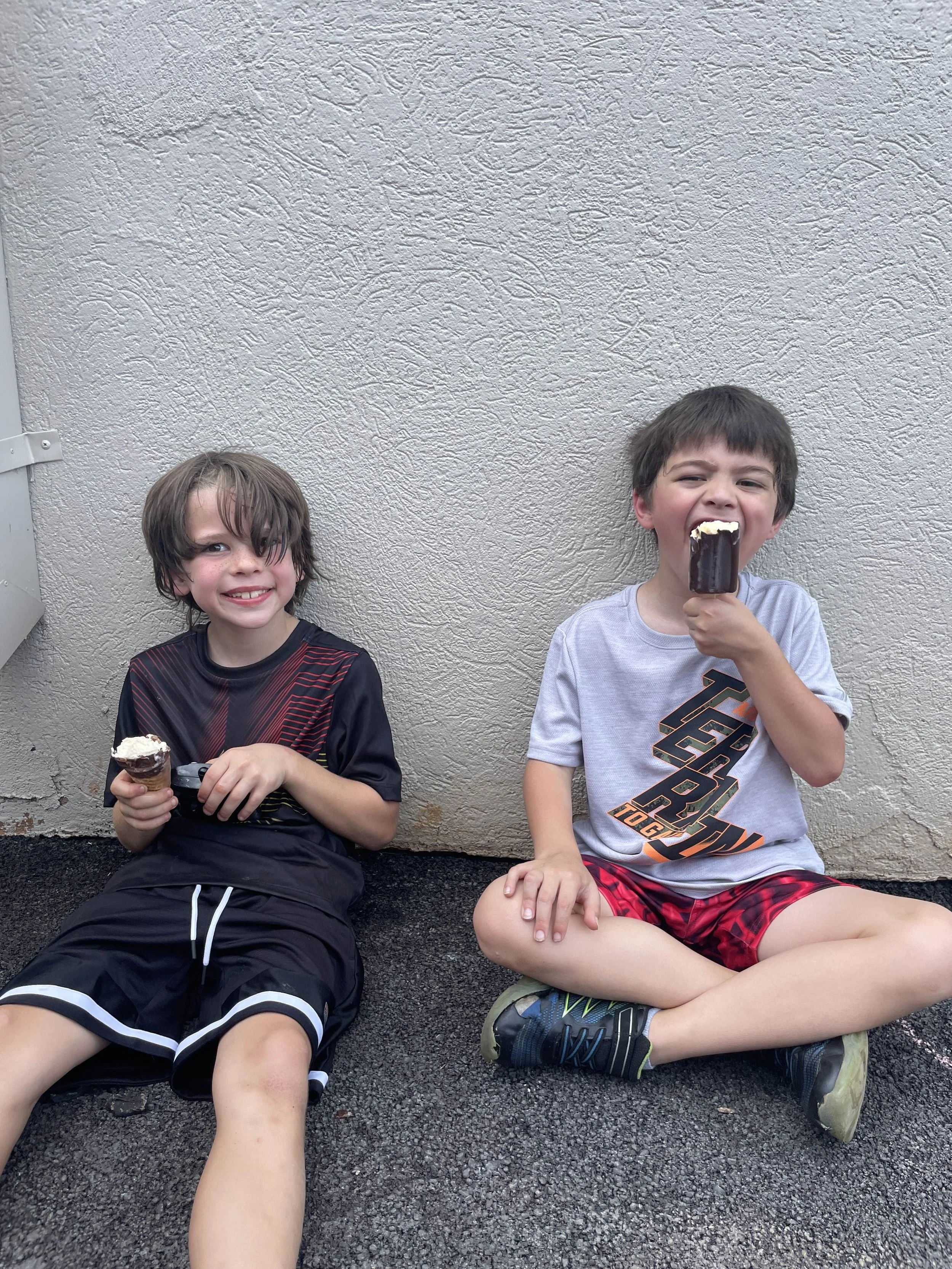 Two young boys sitting on wet pavement against a beige textured wall, enjoying ice cream bars.