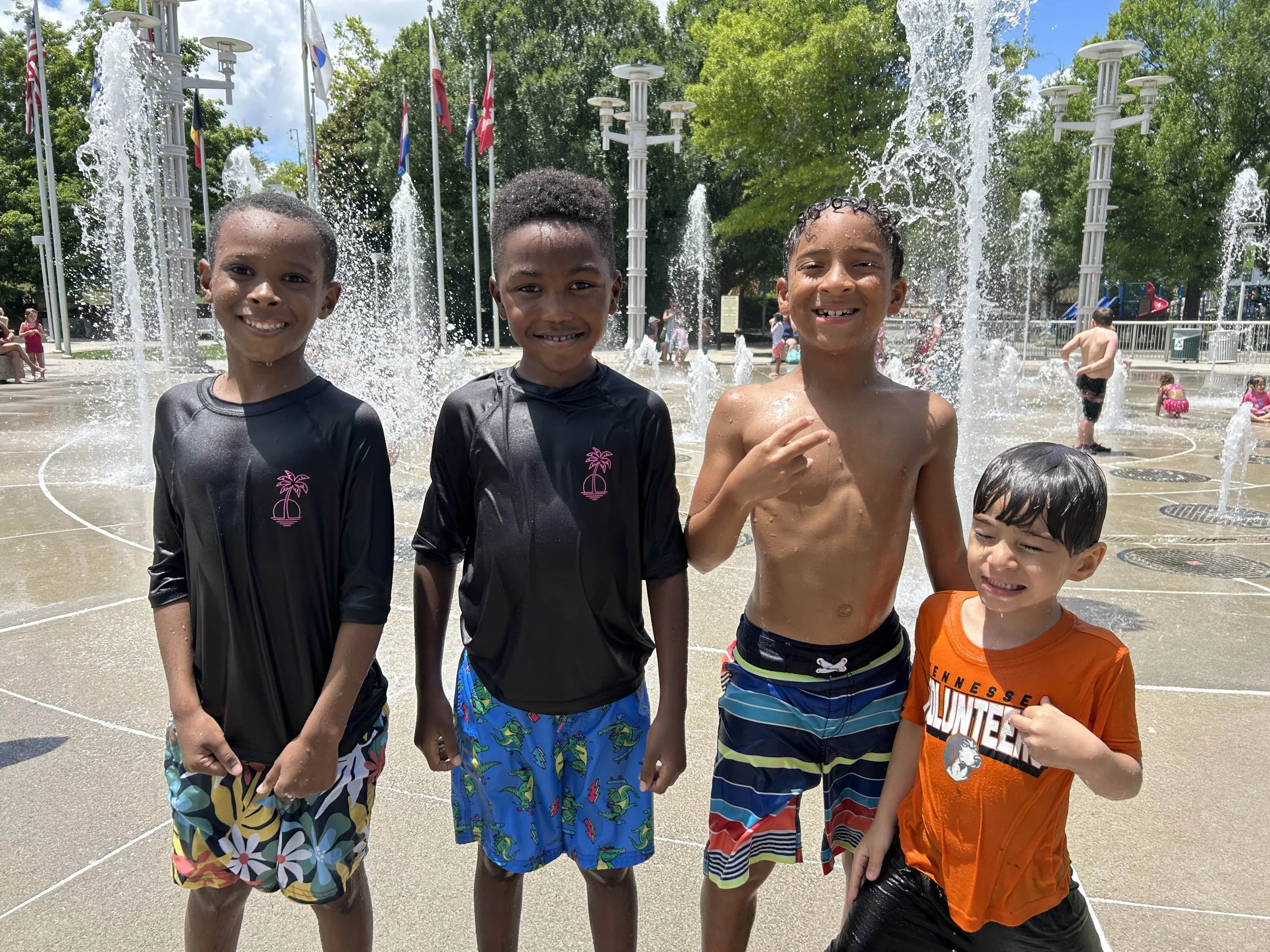 Four boys in swimsuits smiling and posing in front of fountains at a splash pad on a sunny day.