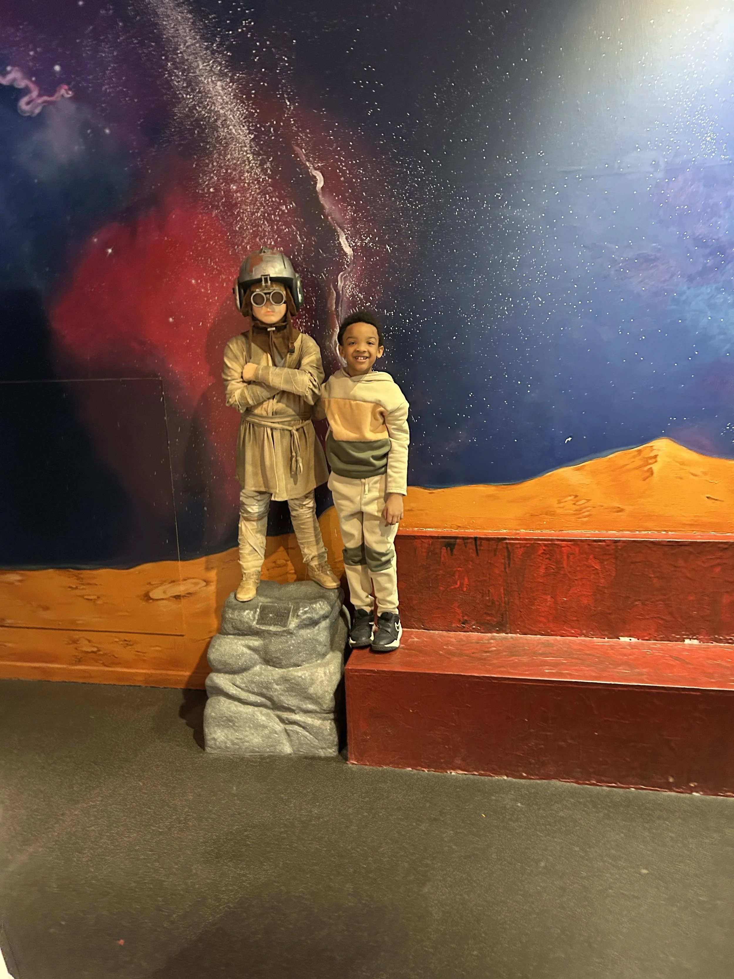 Two children standing inside a space-themed museum exhibit with a painted backdrop of outer space and mountains. One child is dressed as an astronaut with a helmet and goggles, and the other is smiling and wearing casual clothing.