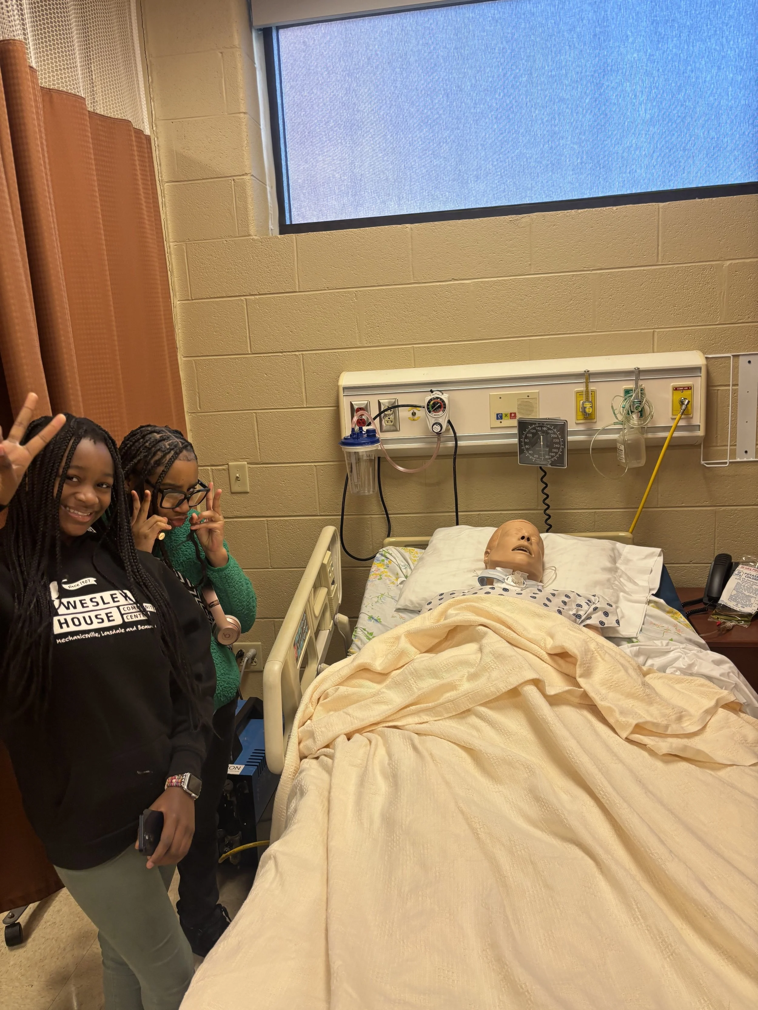 Two young girls standing next to a hospital bed with a medical mannequin on it, posing and smiling. The room has a beige wall, a window, and medical equipment attached to the wall.