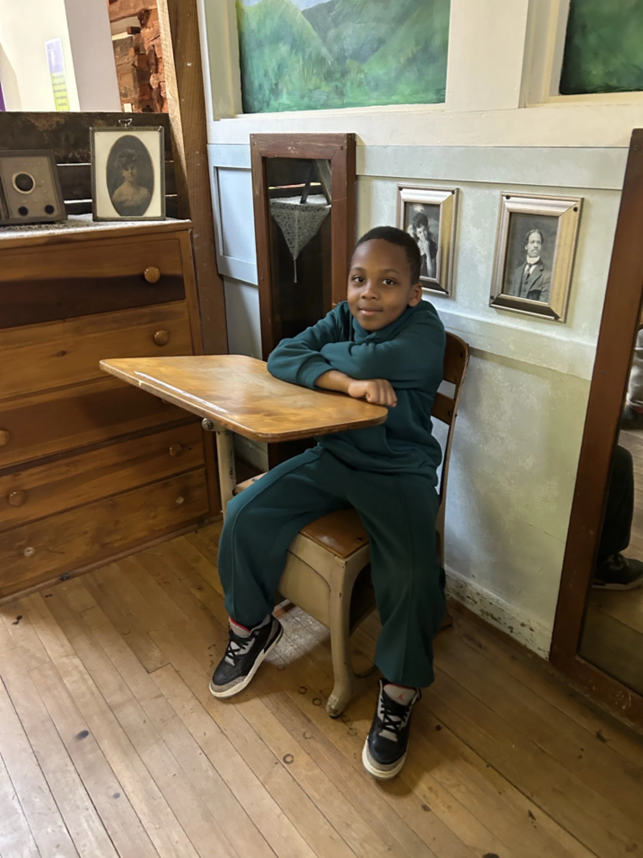 A young boy sitting in a wooden chair at a small table in a vintage room with framed photographs and paintings on the walls.