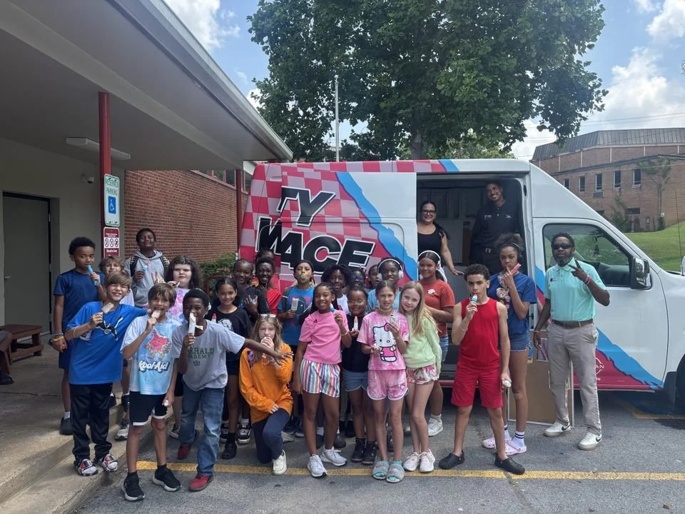 Group of children and adults standing in front of a Y MACE ice cream truck, some children holding ice cream cones, outdoors on a cloudy day with a large tree in the background.