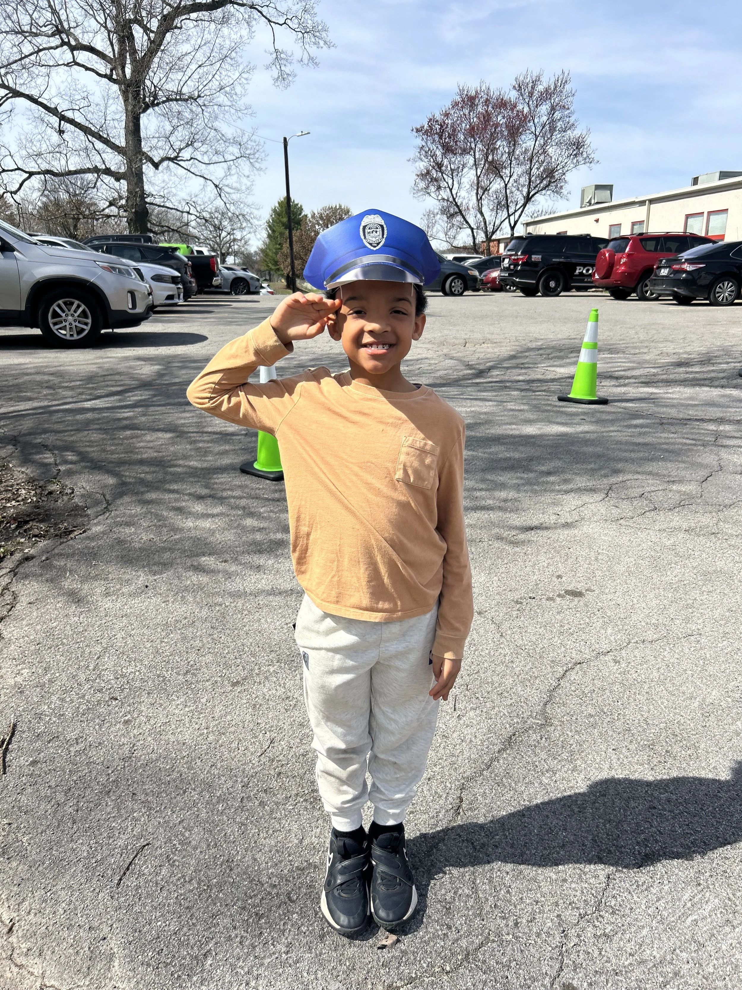 A young boy wearing a blue police officer's hat, pink shirt, and beige pants, saluting and smiling in a parking lot with cars and orange traffic cones.