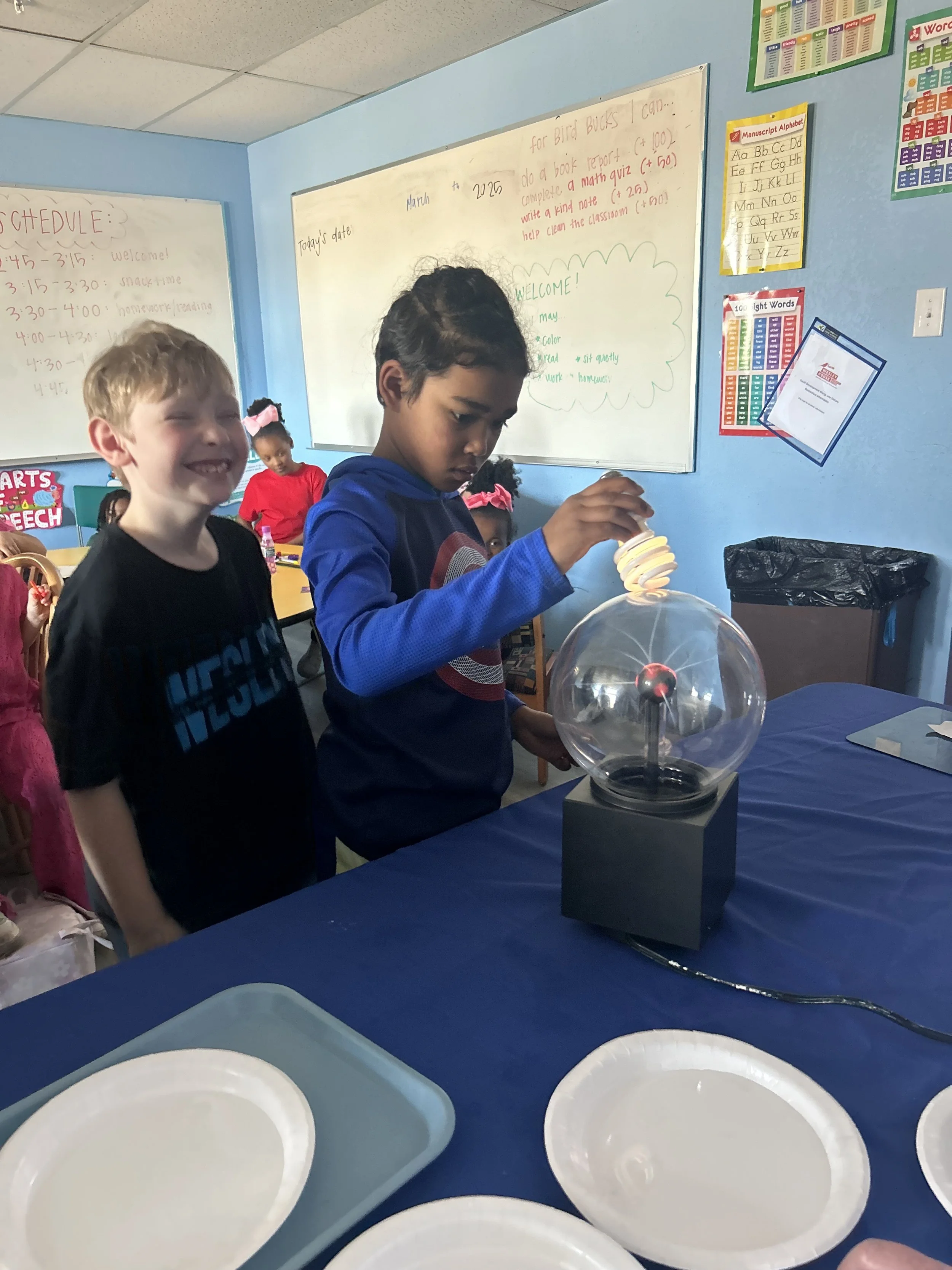 Children in a classroom experiment with a Tesla coil that produces sparks, and others watching with smiles.